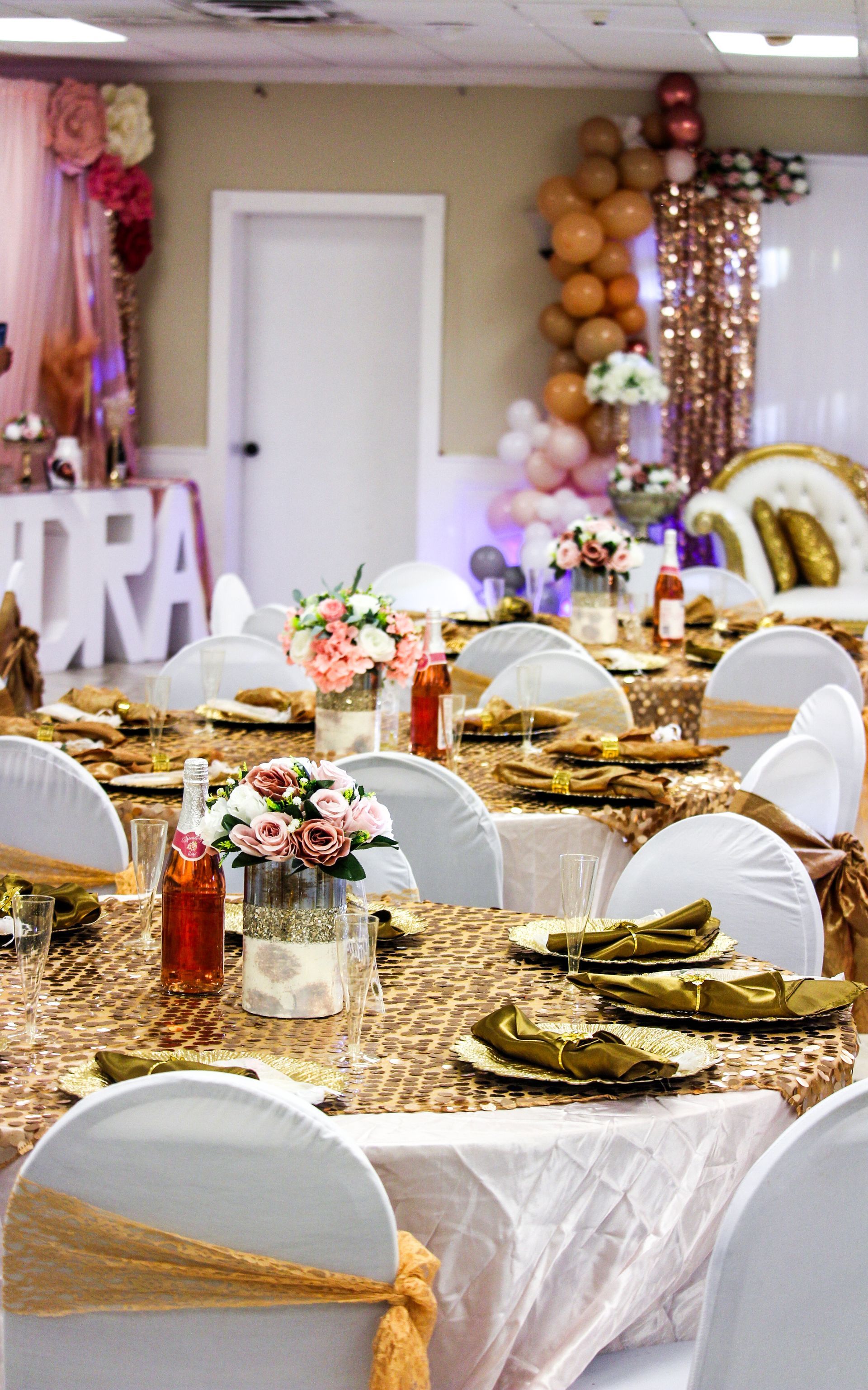 Decorated banquet table with gold accents, white chair covers, and pink balloon arch at an event venue