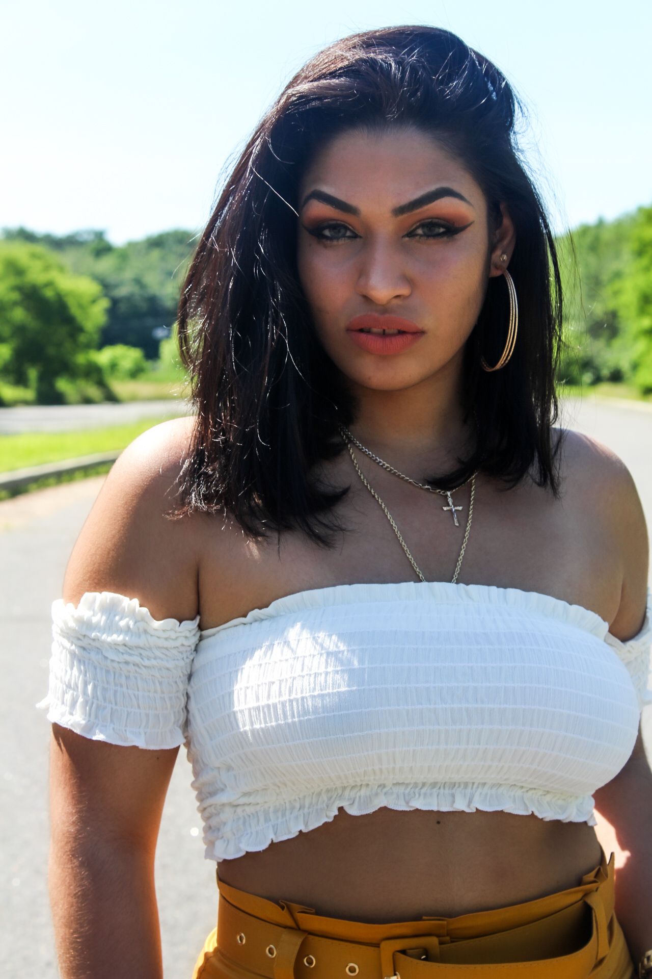 Woman standing outdoors in a white off-shoulder top and tan belt, with a green park background.