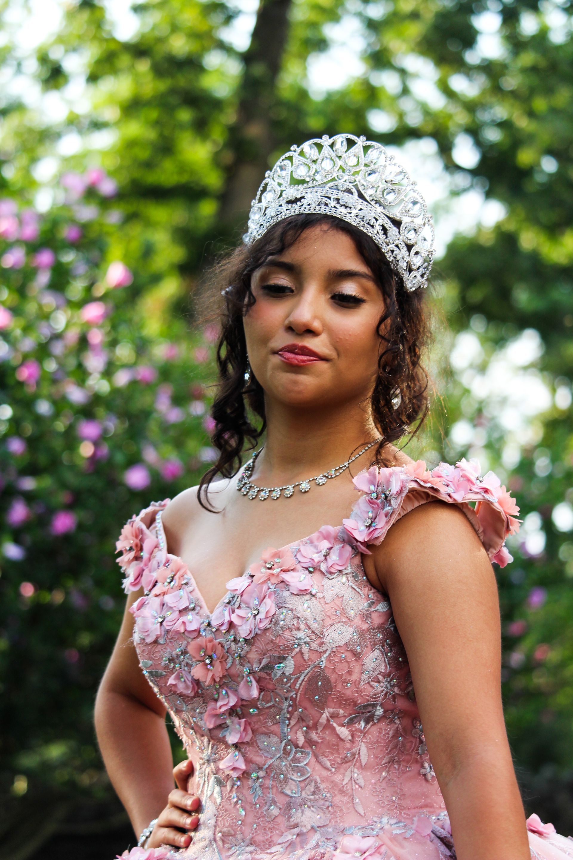 Woman in a pink floral gown and jeweled tiara, posing outdoors with greenery and flowers behind her.