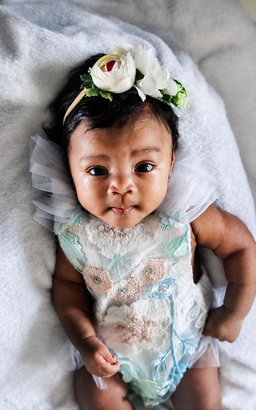 Infant in a white dress with a floral headband, lying on a white blanket and looking up at the camera