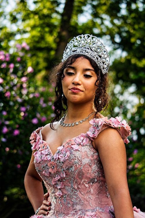 Woman in a pink floral dress and jeweled crown posing outdoors against a blurred garden backdrop