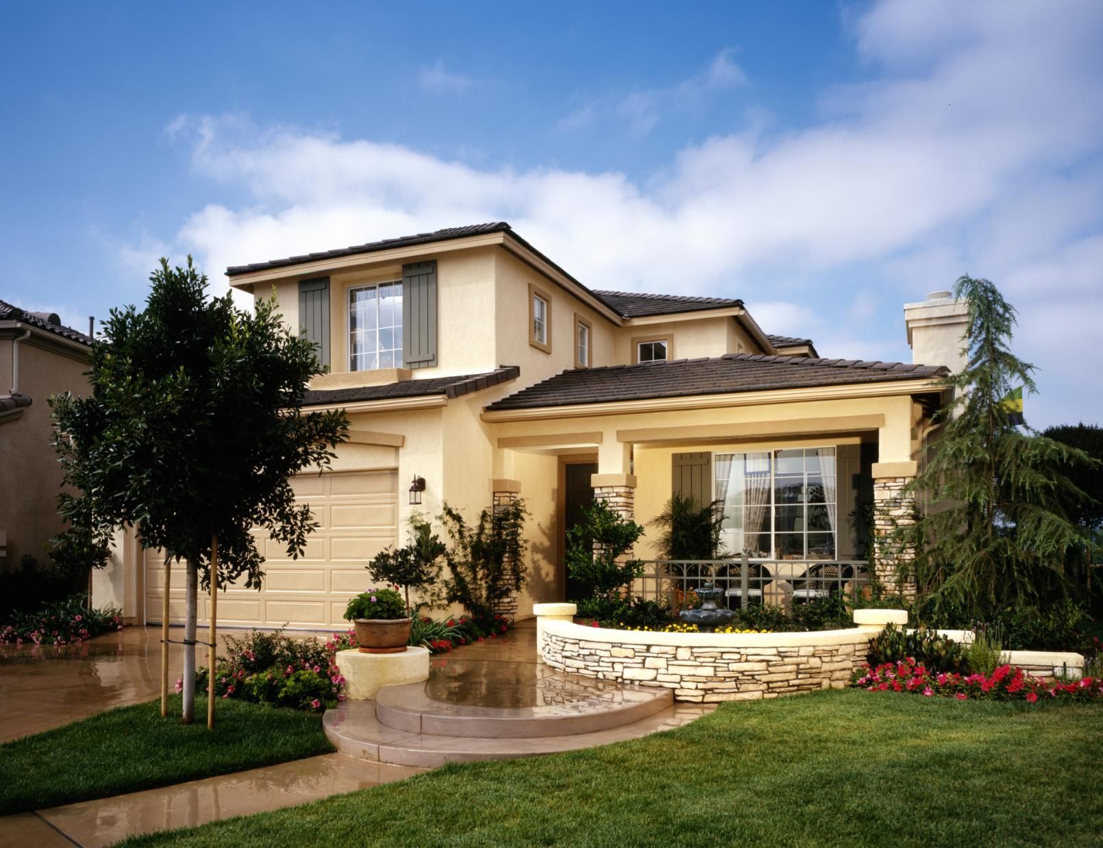 Two-story beige suburban house with a manicured lawn, patio, and landscaped front yard