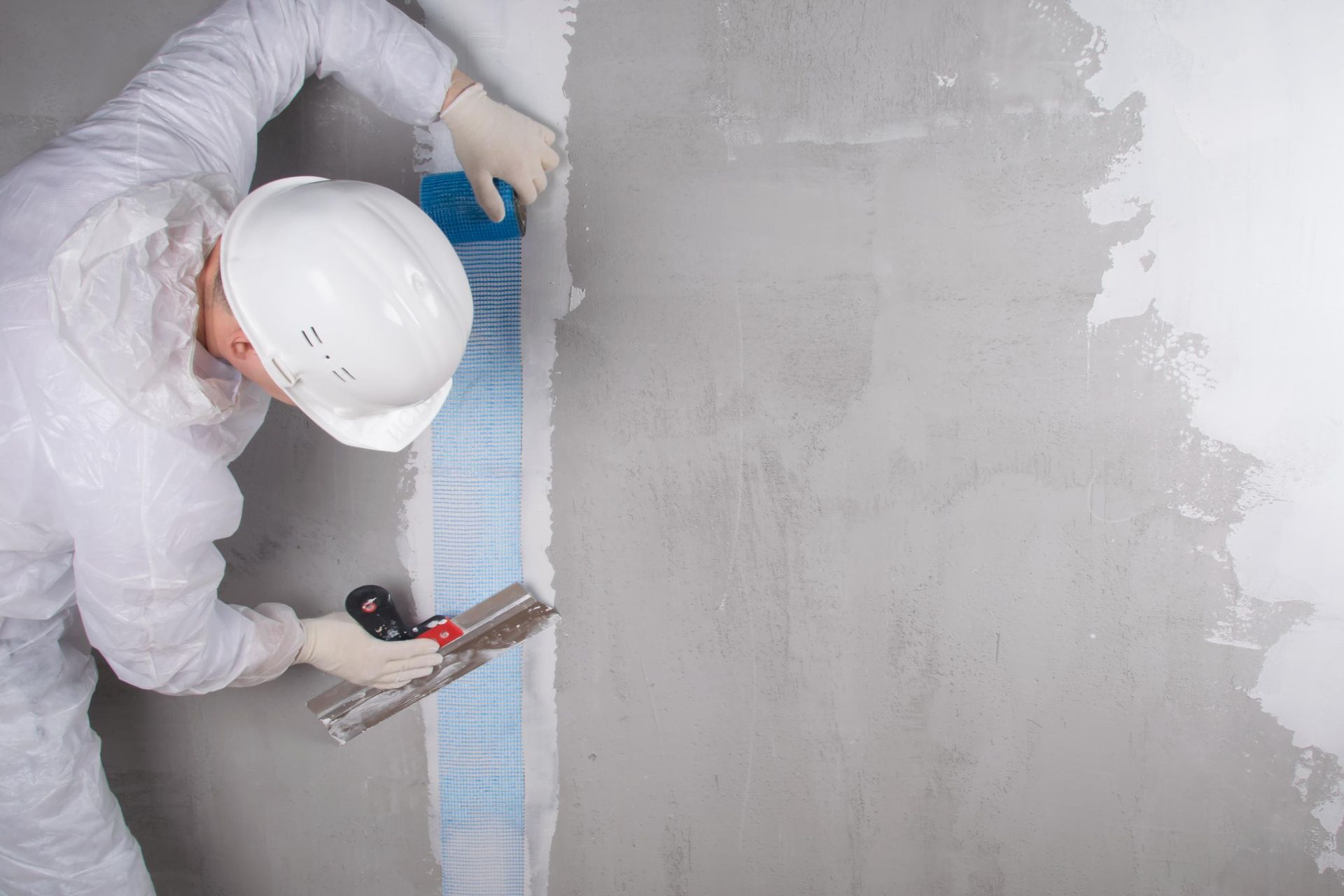 Worker in white coveralls troweling plaster onto a wall beside blue mesh tape