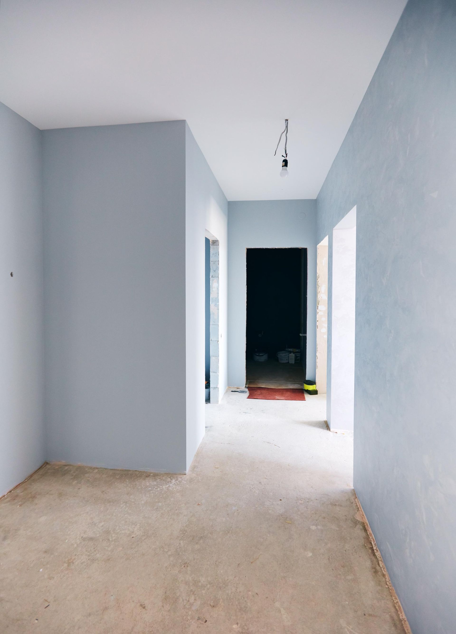 Empty hallway with light blue walls, white ceiling, and unfinished floor leading to a dark doorway.