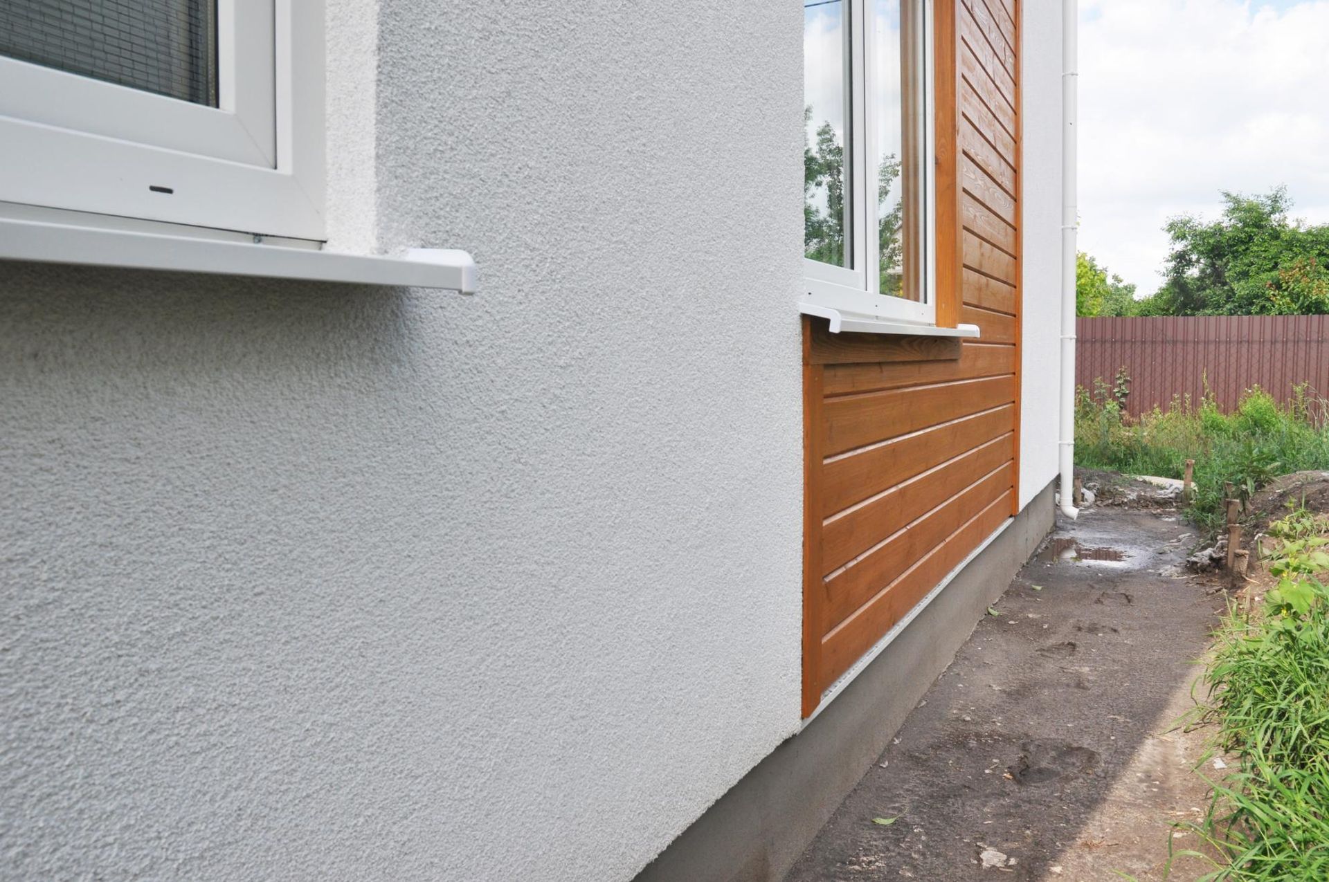 Side yard beside a house with gray stucco, wood siding, and a narrow dirt path by a fence.