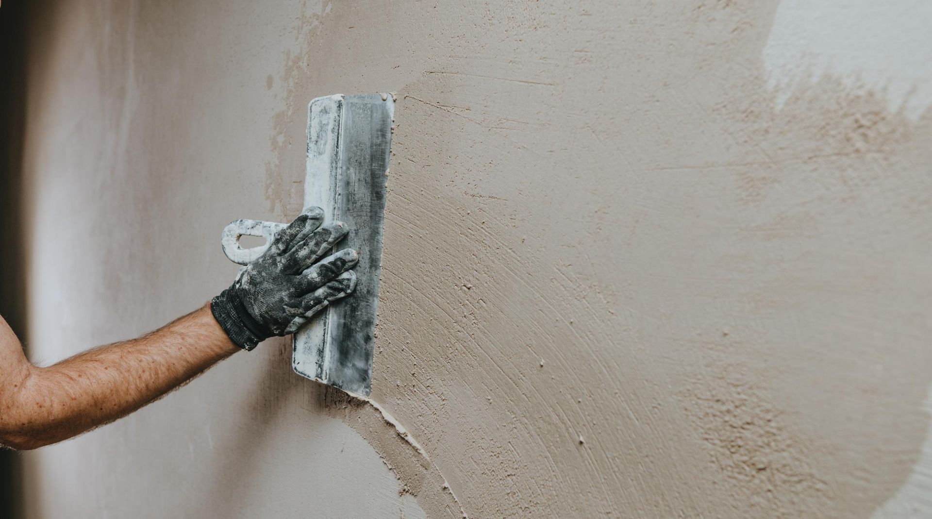 A gloved hand smooths wet plaster on a beige wall with a trowel.