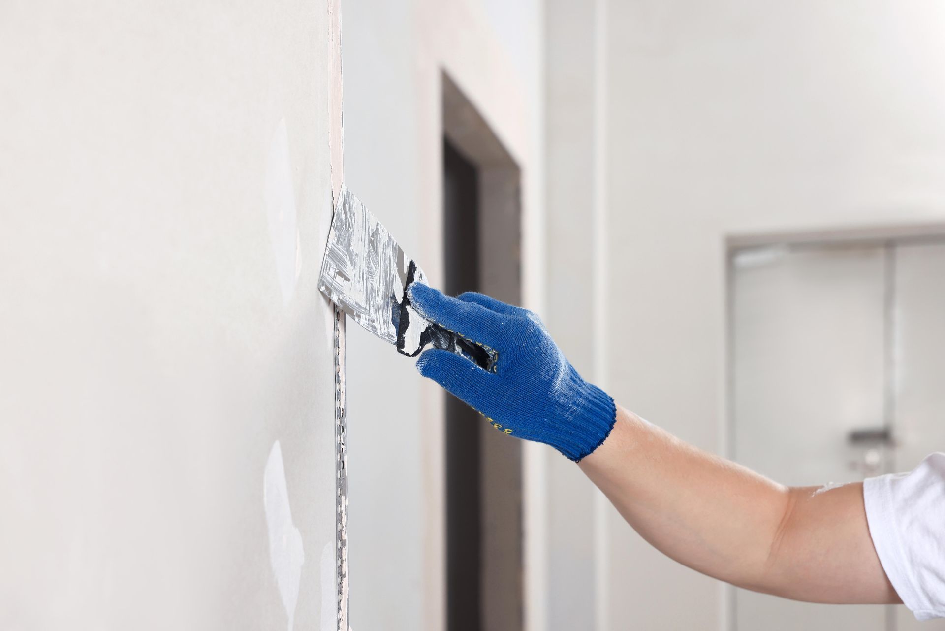 Gloved hand applying white paint or filler to a wall with a putty knife near a doorway