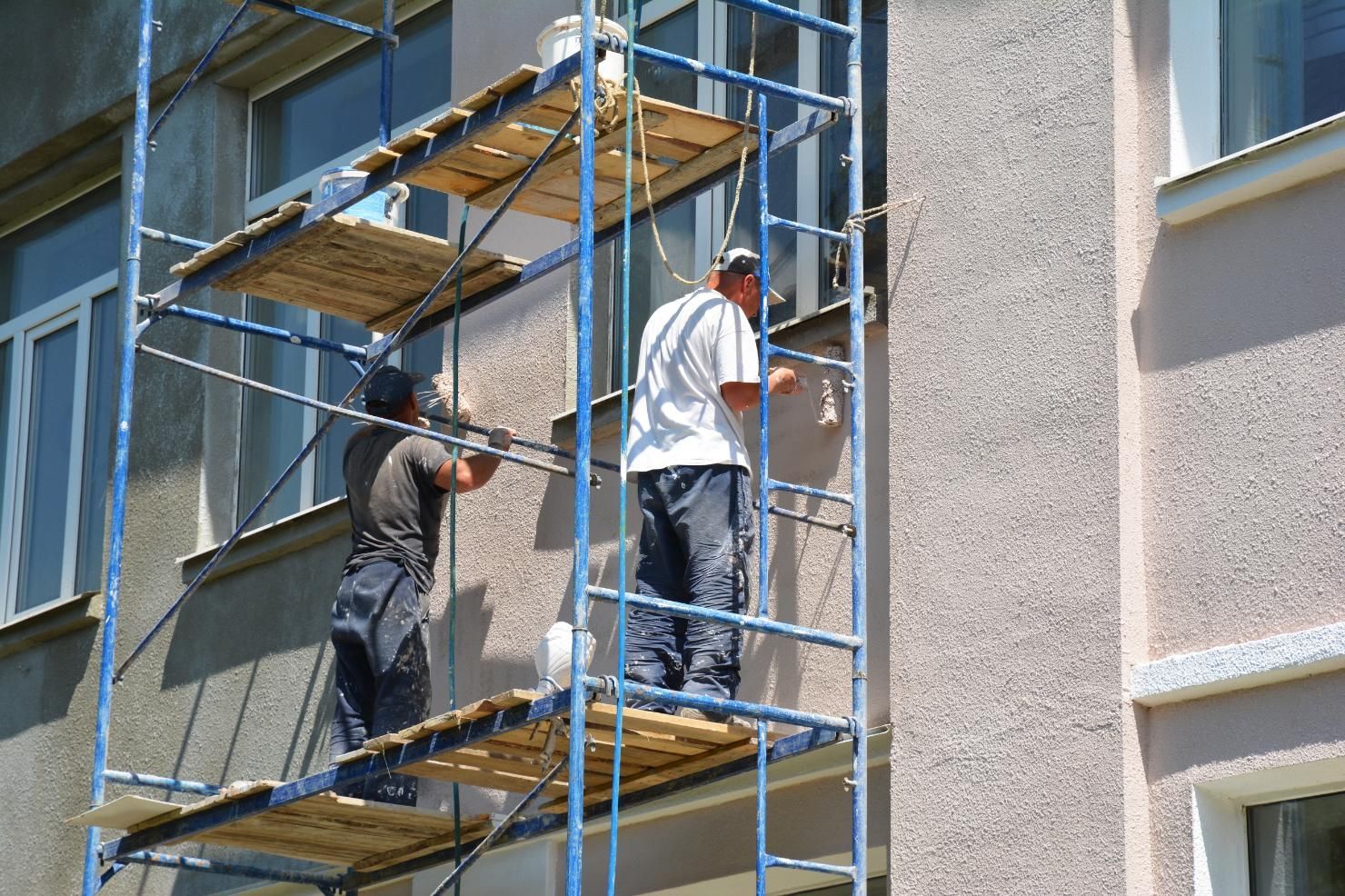 Two workers on scaffolding painting a building exterior next to windows