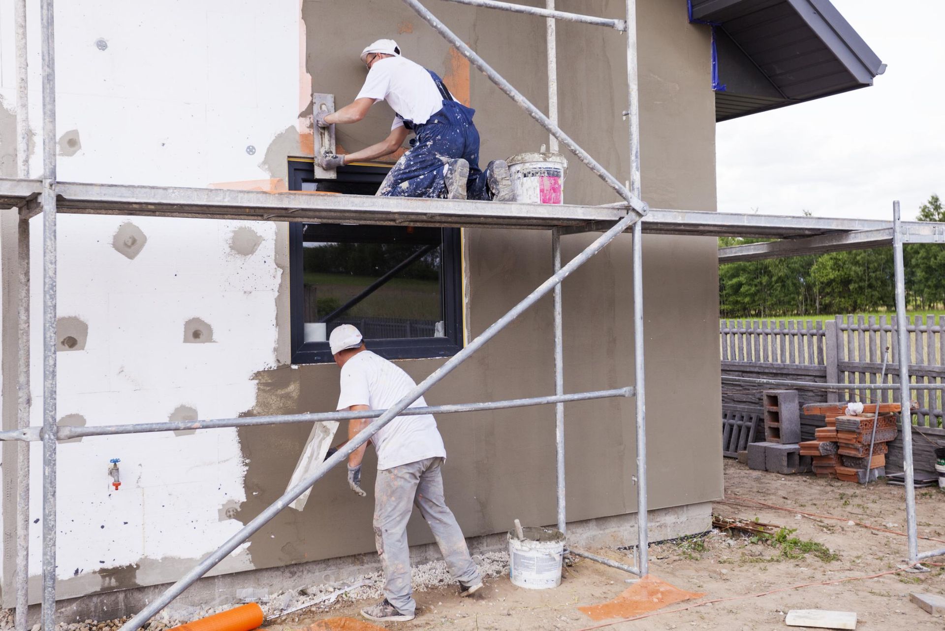 Two workers applying exterior stucco to a house wall with scaffolding and buckets nearby