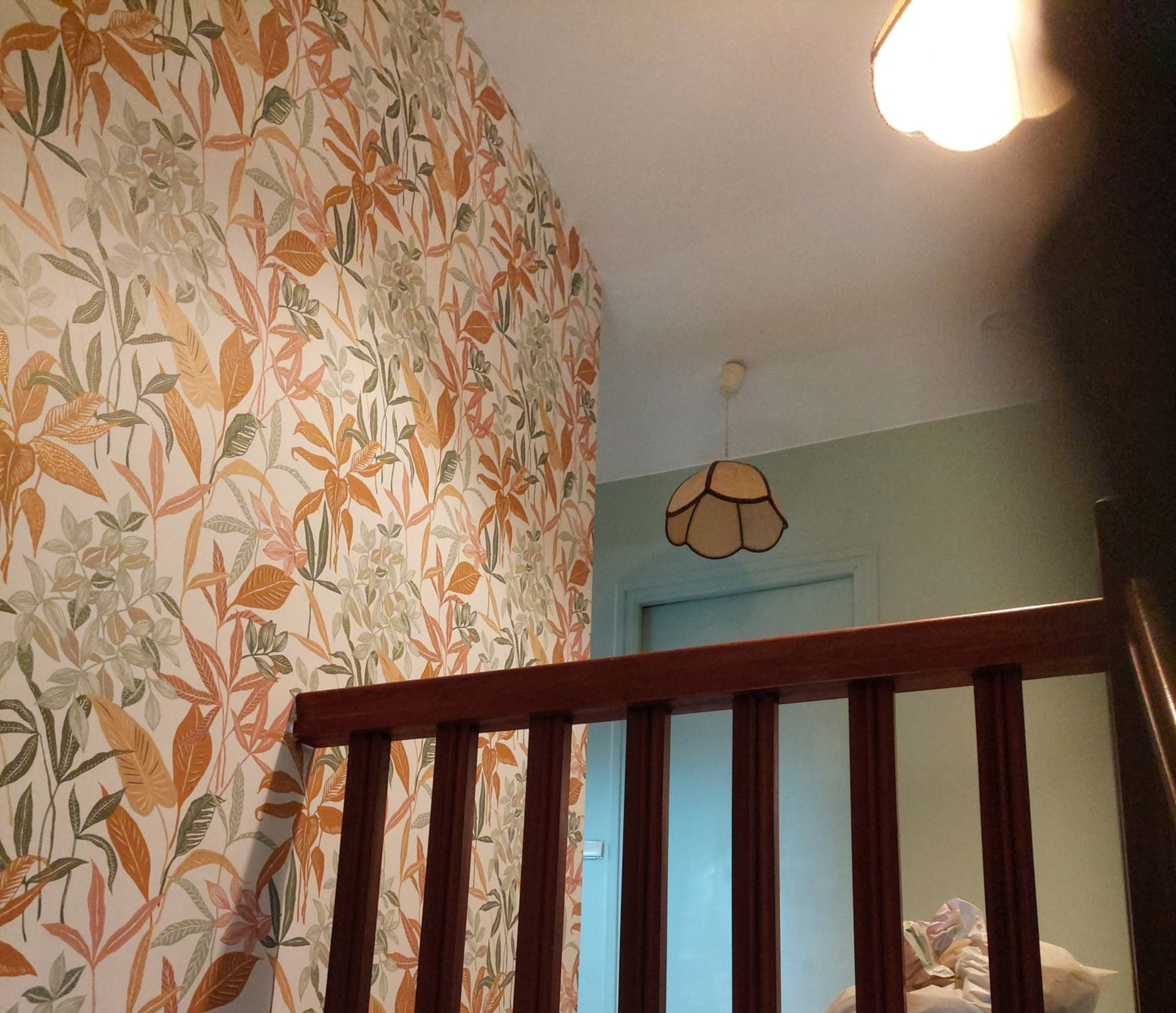 Cage d’escalier avec du papier peint fleuri, une rampe en bois, un mur bleu pâle et un plafonnier suspendu.
