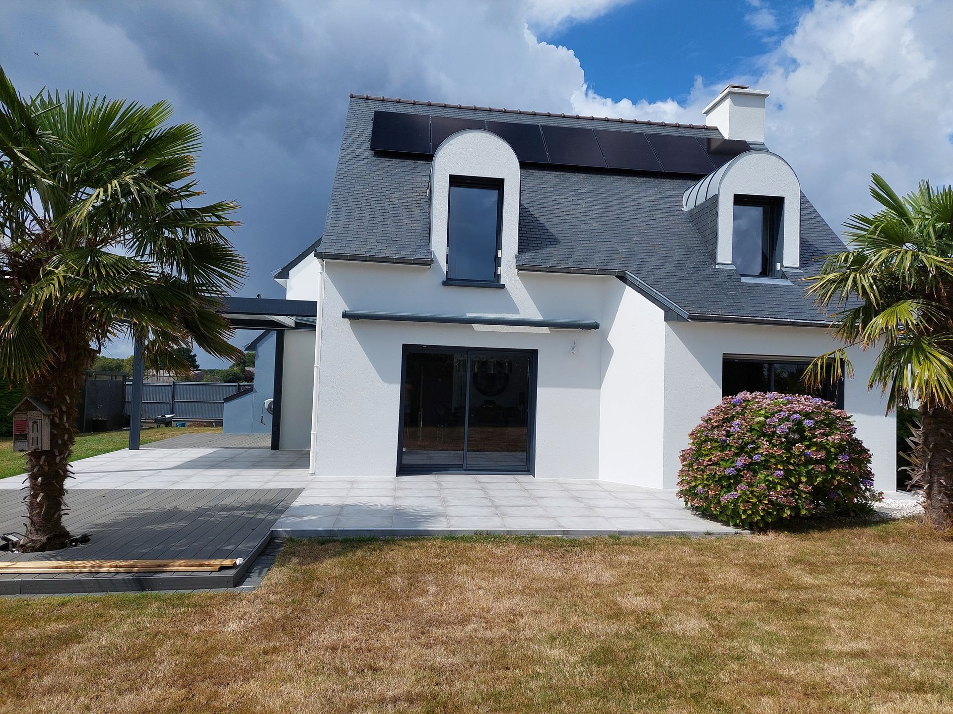 Maison blanche au toit d’ardoise foncé, avec un porche, un palmier et une pelouse sèche sous un ciel bleu.