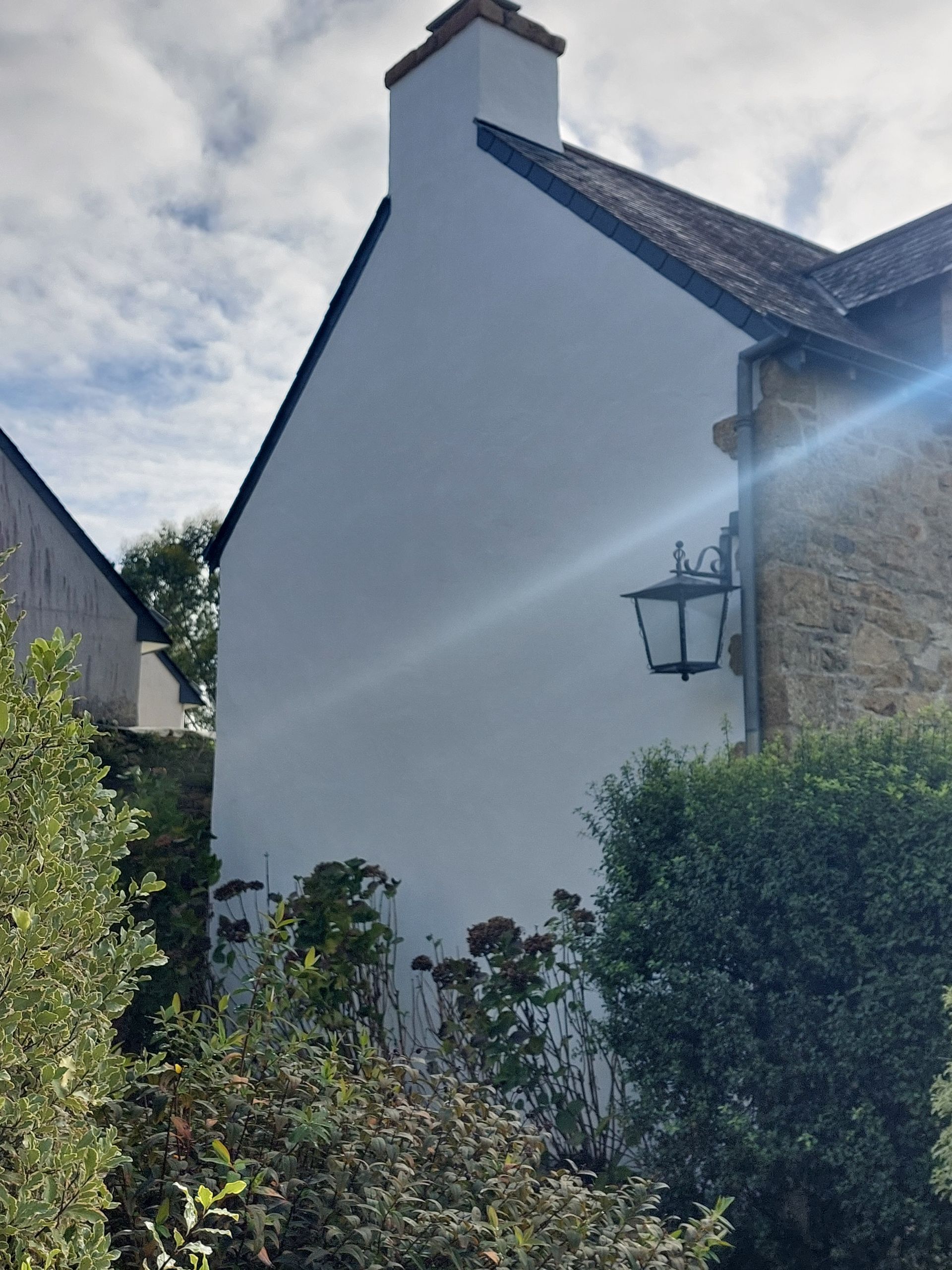 Mur latéral blanc d’une maison, avec une lanterne noire, des haies et un éclat de soleil vif sous un ciel nuageux