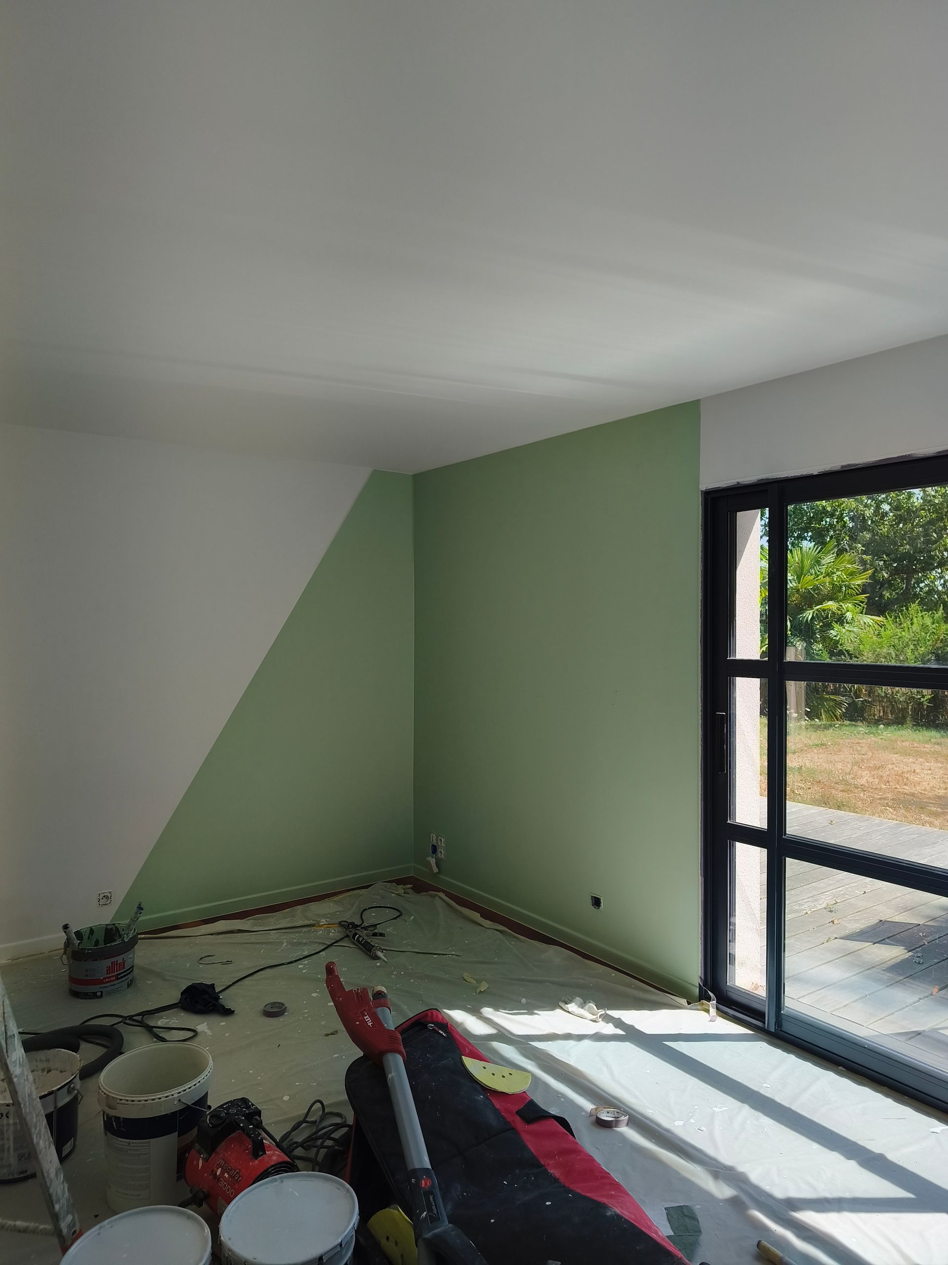 Pièce partiellement peinte, avec un mur d’accent vert, des outils au sol et la lumière du soleil qui passe par une grande porte coulissante