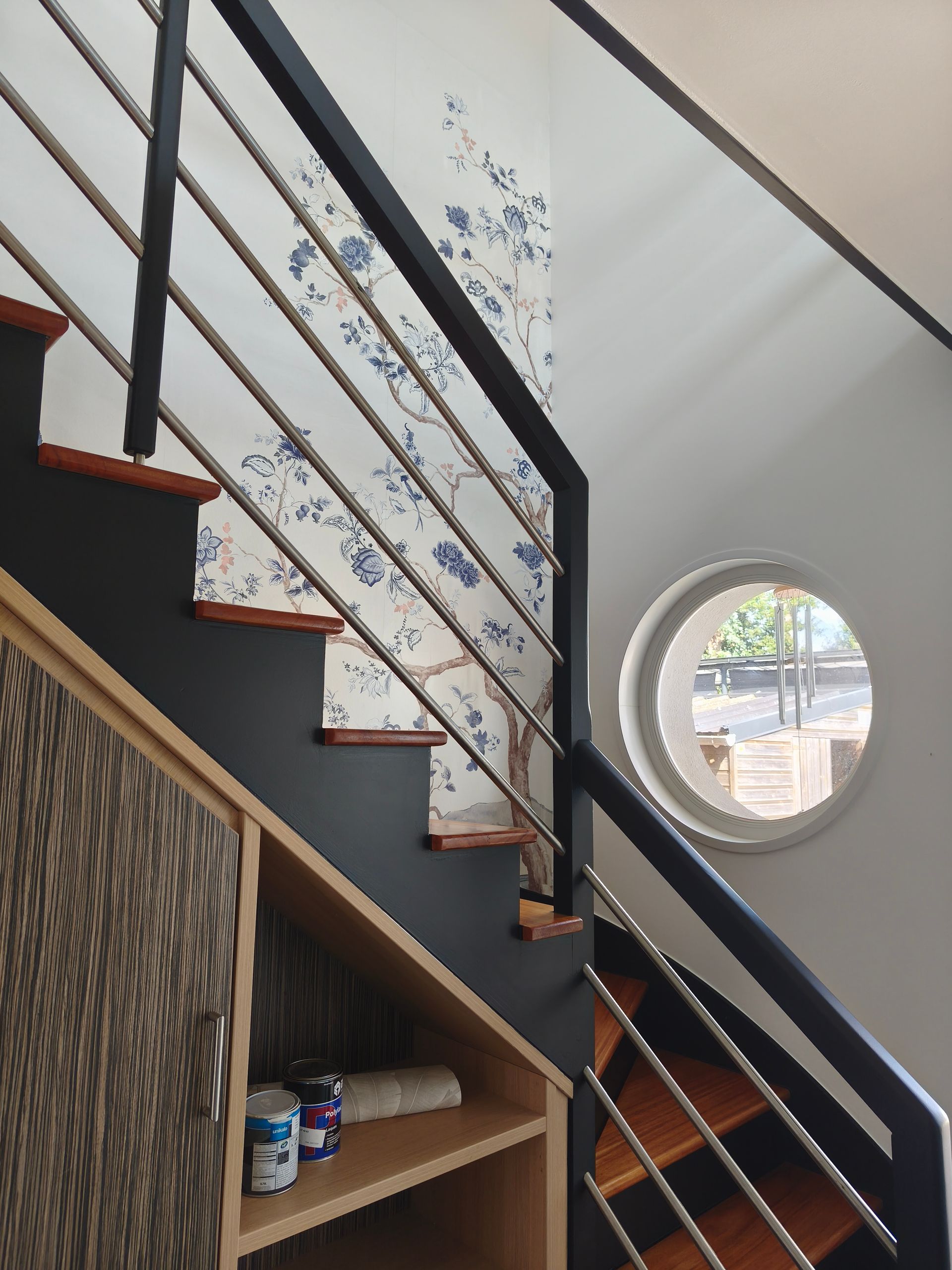Escalier avec des rampes sombres, une fenêtre circulaire et un mur à motifs dans un intérieur lumineux.