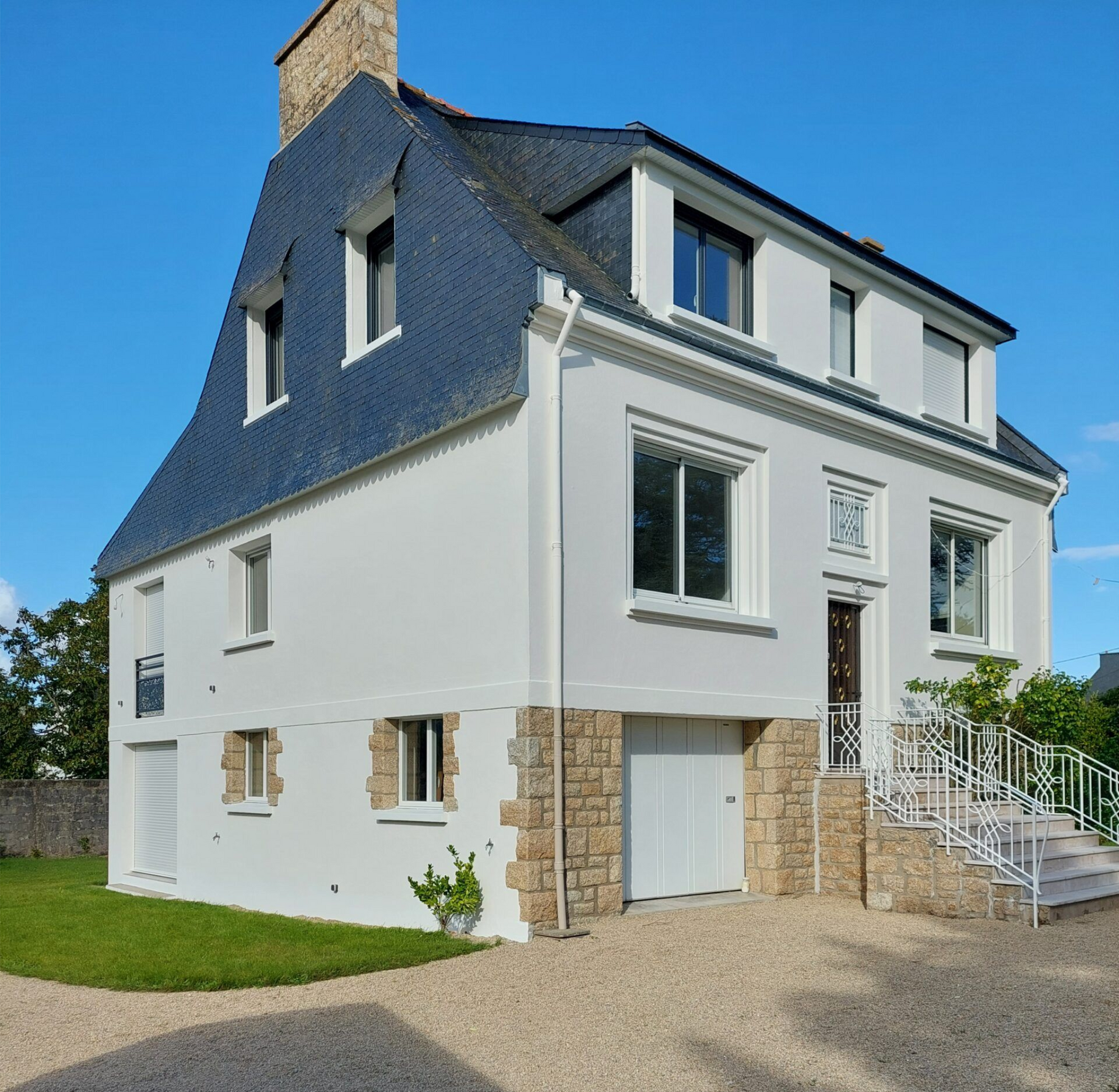 Maison blanche à deux étages avec un toit bleu et des éléments en pierre, vue depuis le jardin de devant