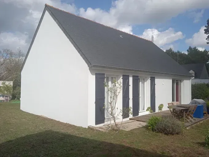 Maison blanche au toit gris pentu et aux portes-fenêtres, vue depuis un jardin enherbé sous un ciel nuageux