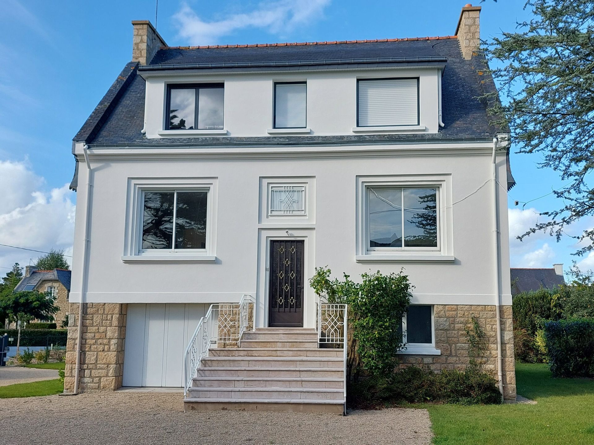 Vue de face d’une maison blanche à deux étages, avec des finitions en pierre, un escalier central et une porte d’entrée foncée
