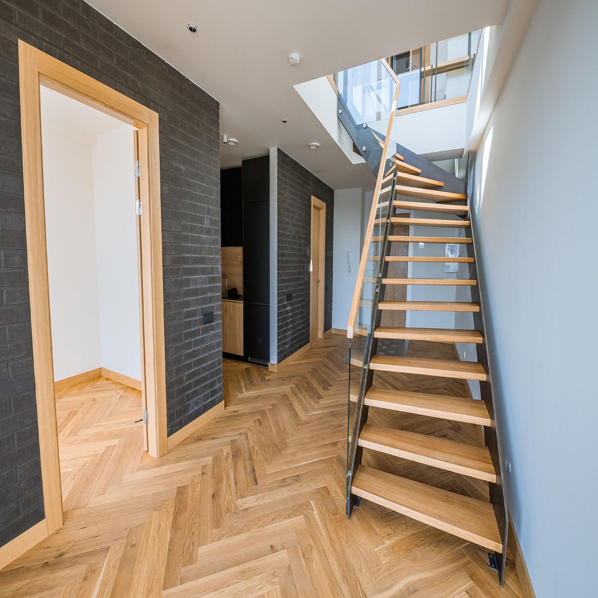 Couloir moderne avec parquet en chevrons, murs en briques sombres et escalier menant à l’étage supérieur
