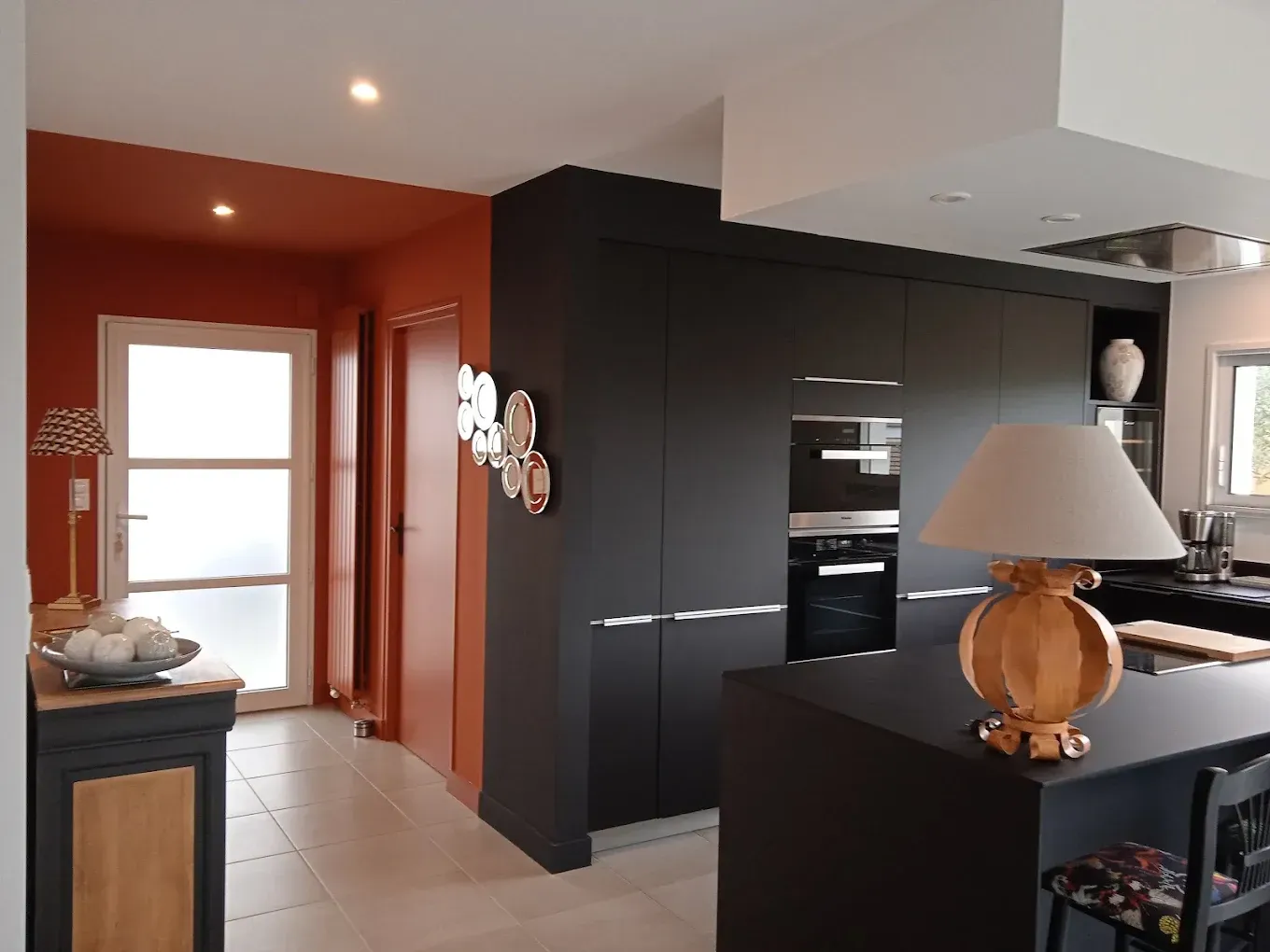 Cuisine moderne avec des placards foncés, un sol carrelé et des murs orange chaleureux ; lampe posée sur le plan de travail au premier plan.