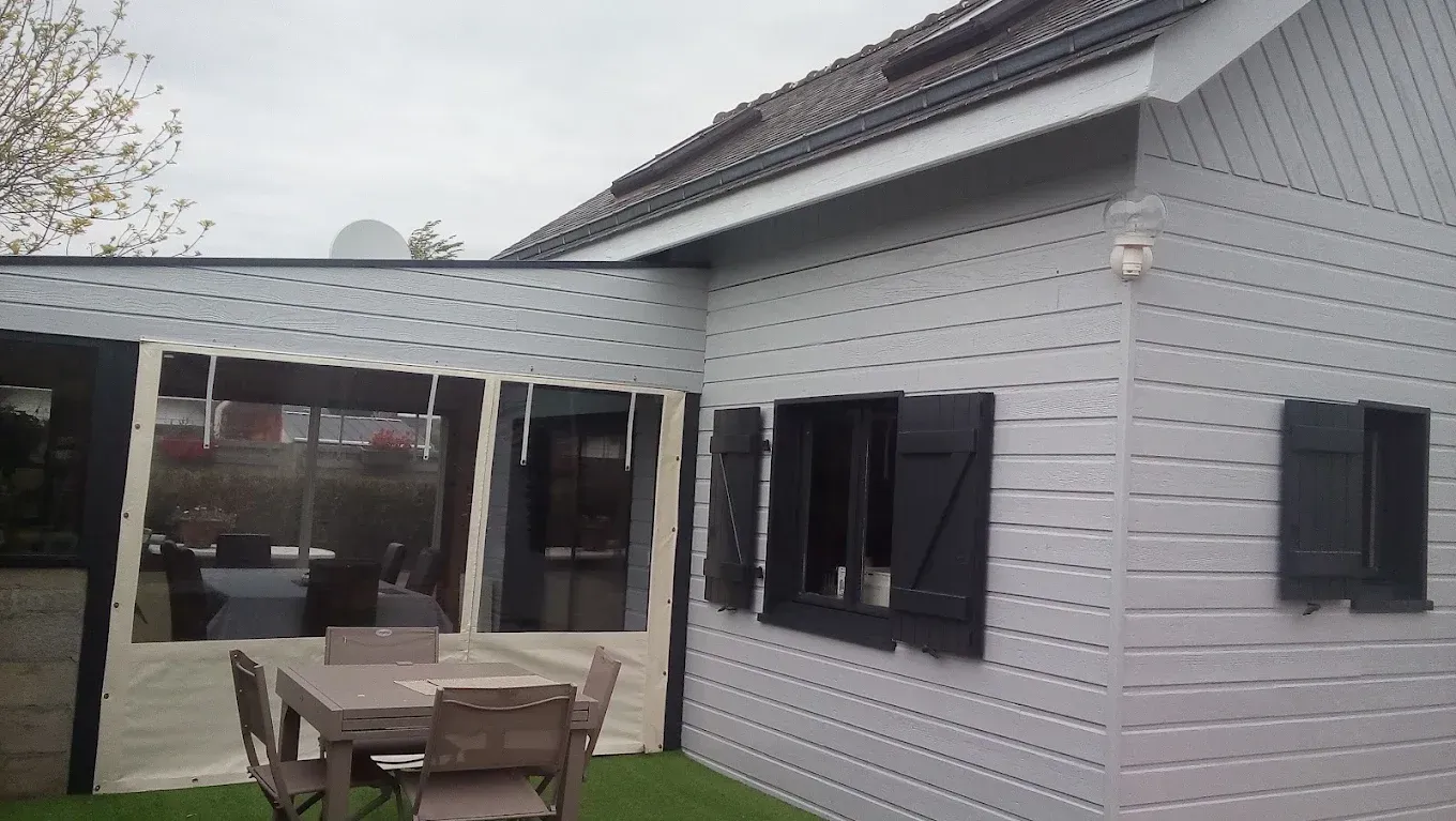 Extérieur de maison blanche avec des volets noirs, une pergola sur la terrasse, ainsi qu’une table et des chaises de jardin