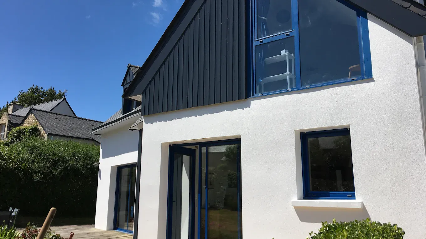 Extérieur d’une maison blanche avec un bardage bleu à l’étage et de grandes portes vitrées sous un ciel dégagé
