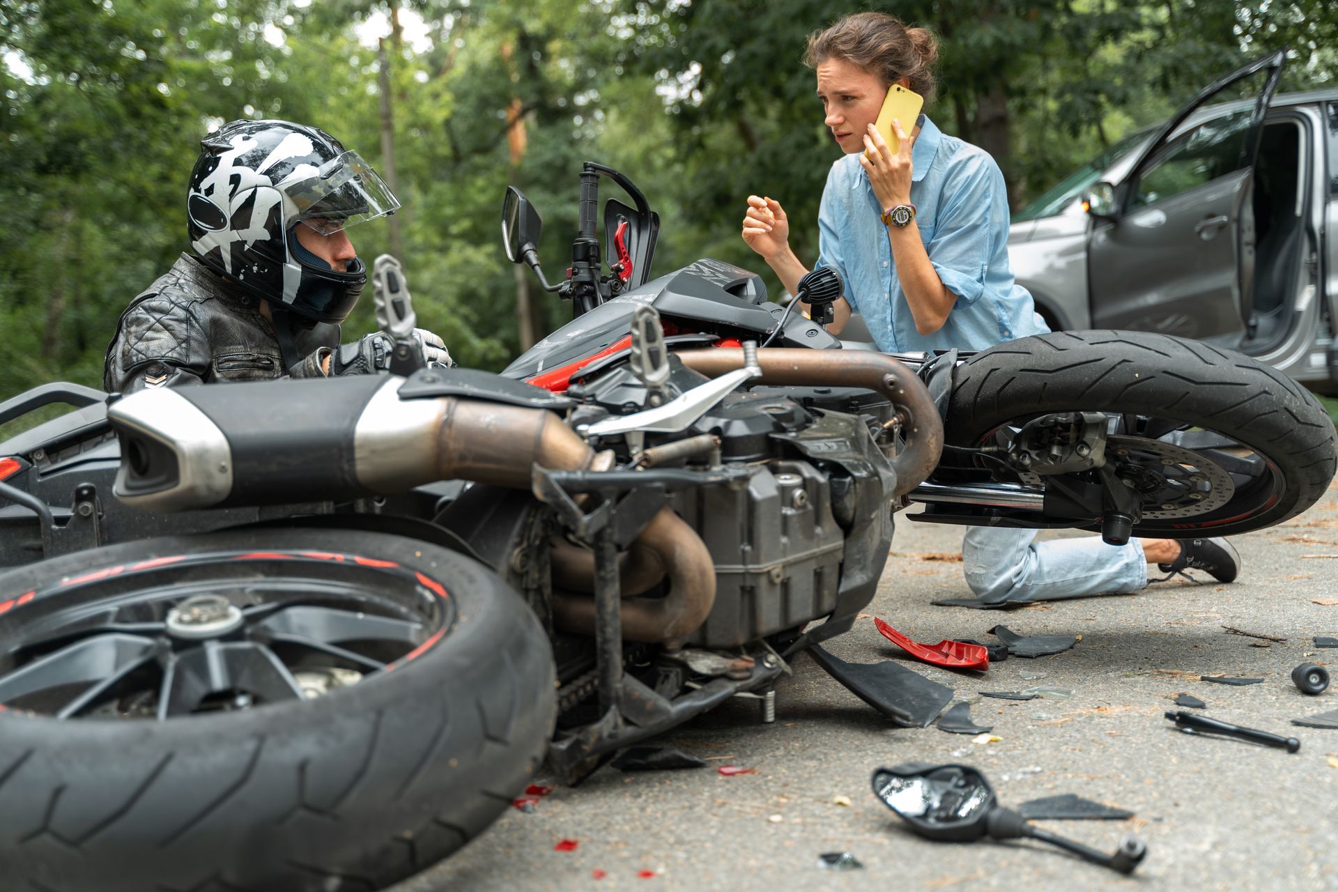 Motorcycle crash scene with overturned bike, debris, and two people talking beside a car and road barrier