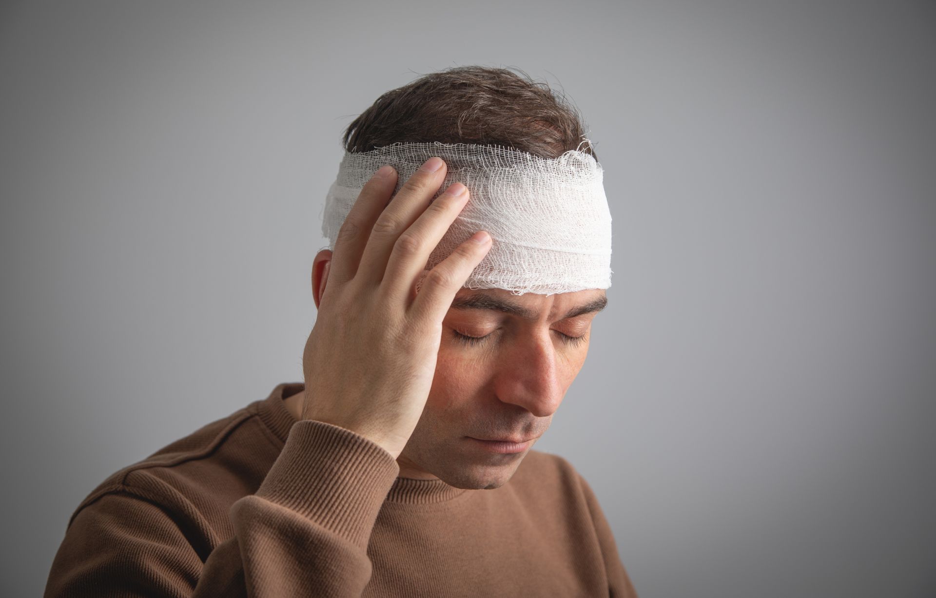 Man with a white head bandage holding his head, looking down against a gray background
