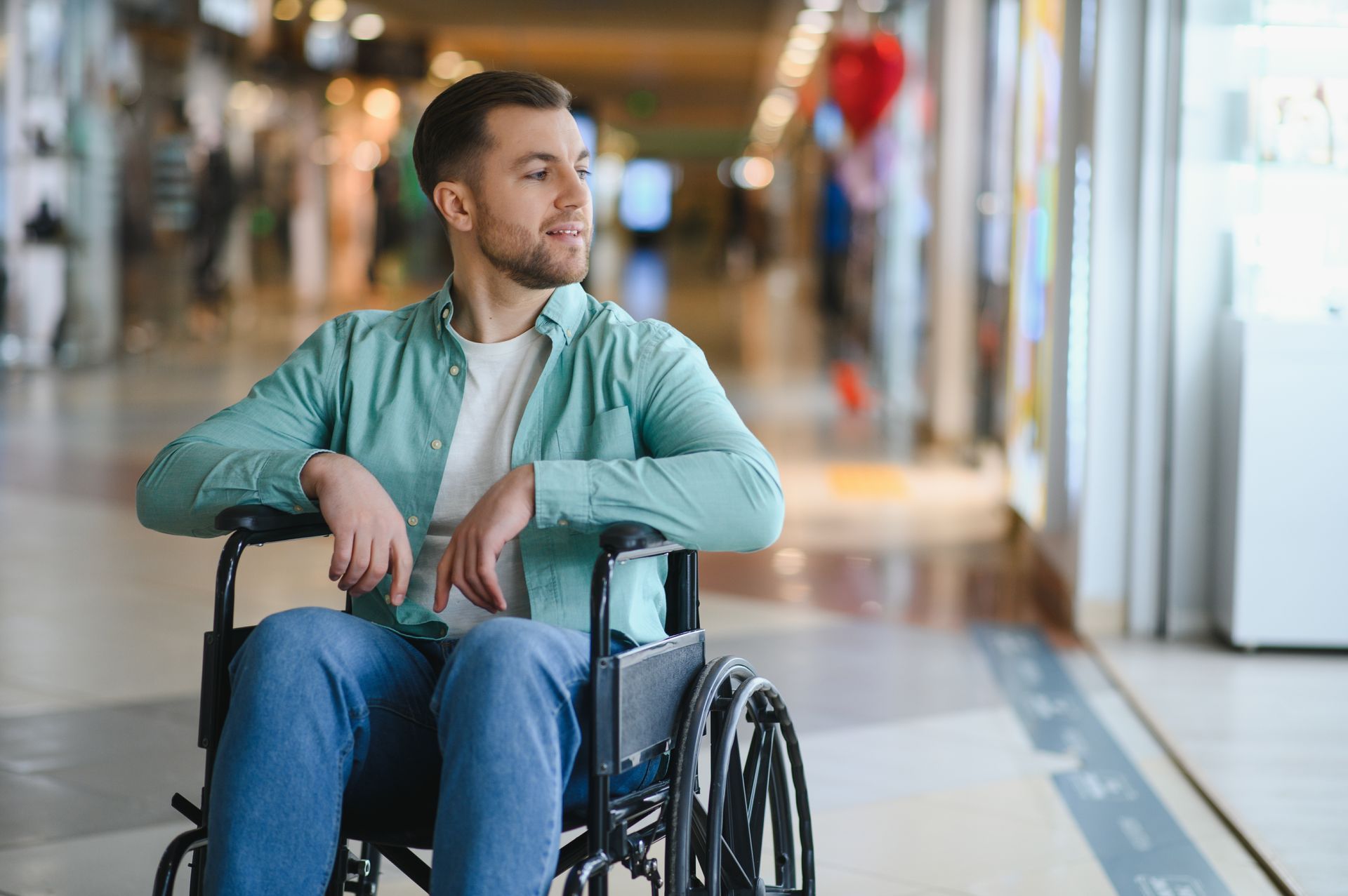 Man in a wheelchair sitting in a bright indoor hallway, looking to the side.