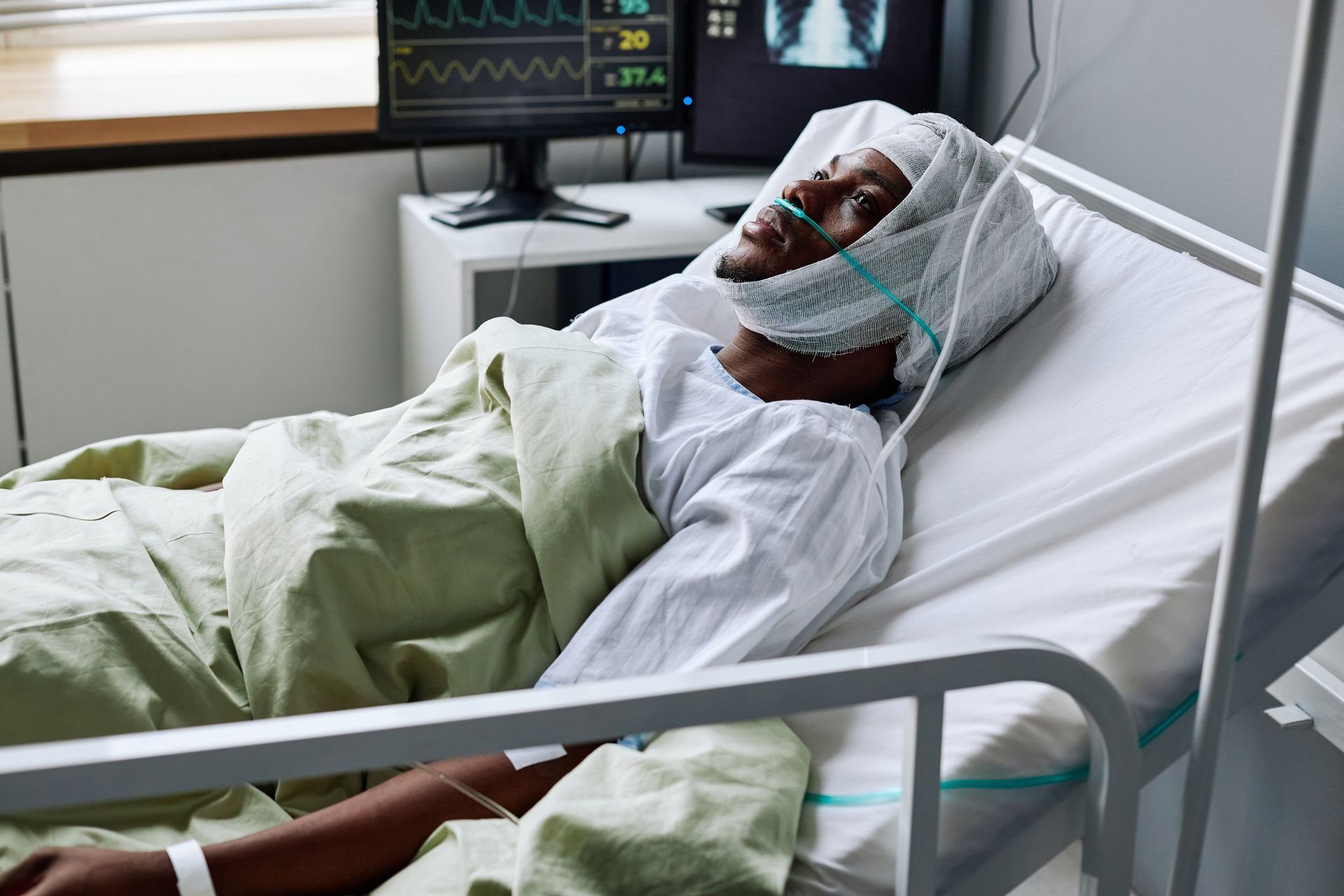Patient lying in a hospital bed, covered with a blanket and wearing a bandaged head