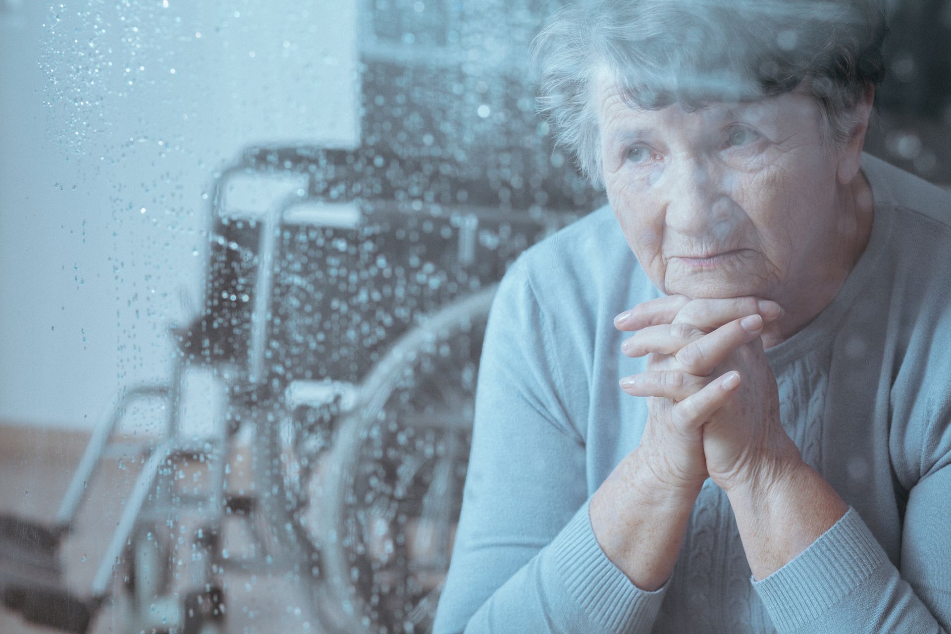Thoughtful person sitting by a rainy window, hands clasped, with a wheelchair in the background.