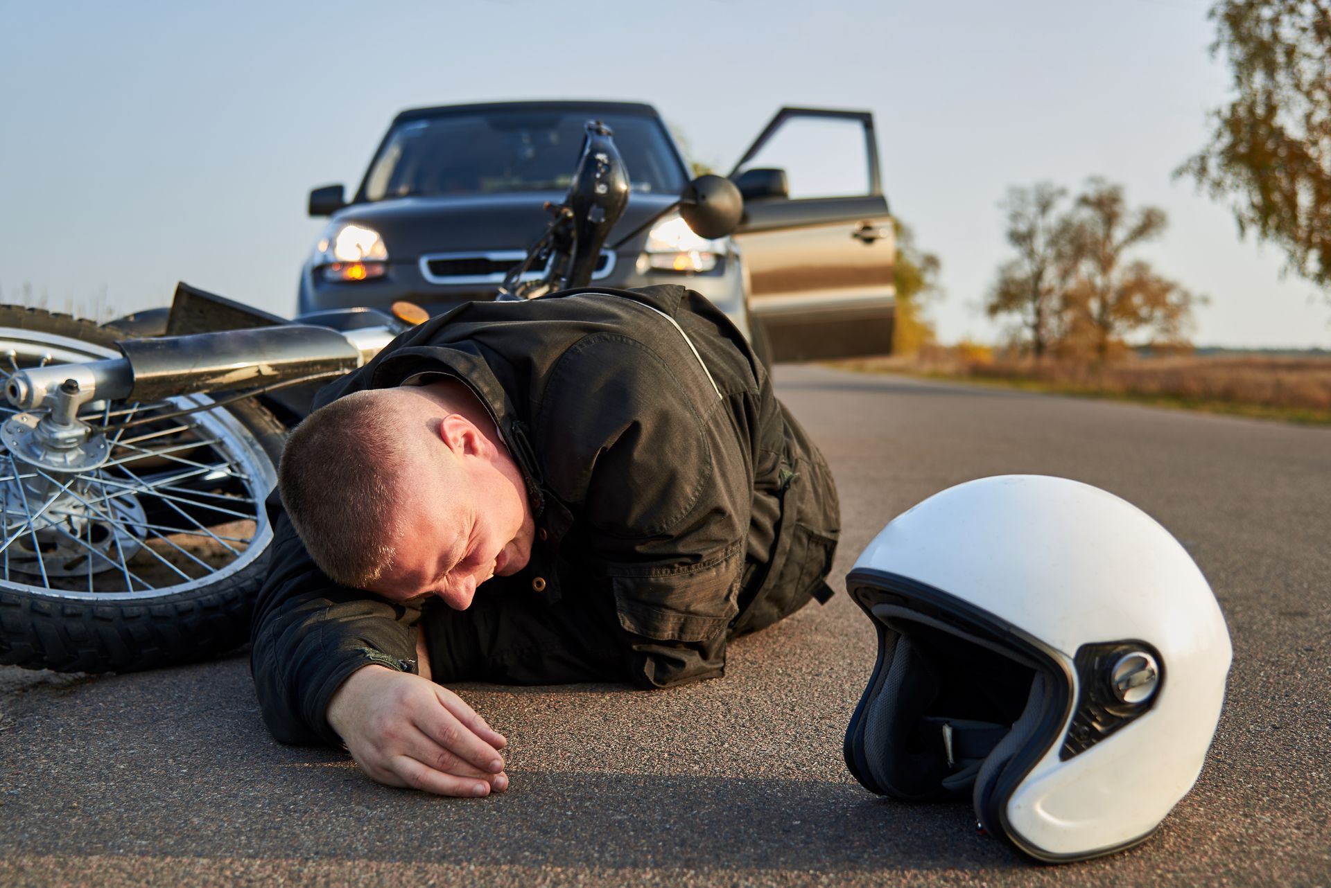 Motorcycle crash scene with injured rider on road, helmet nearby, and car in the background