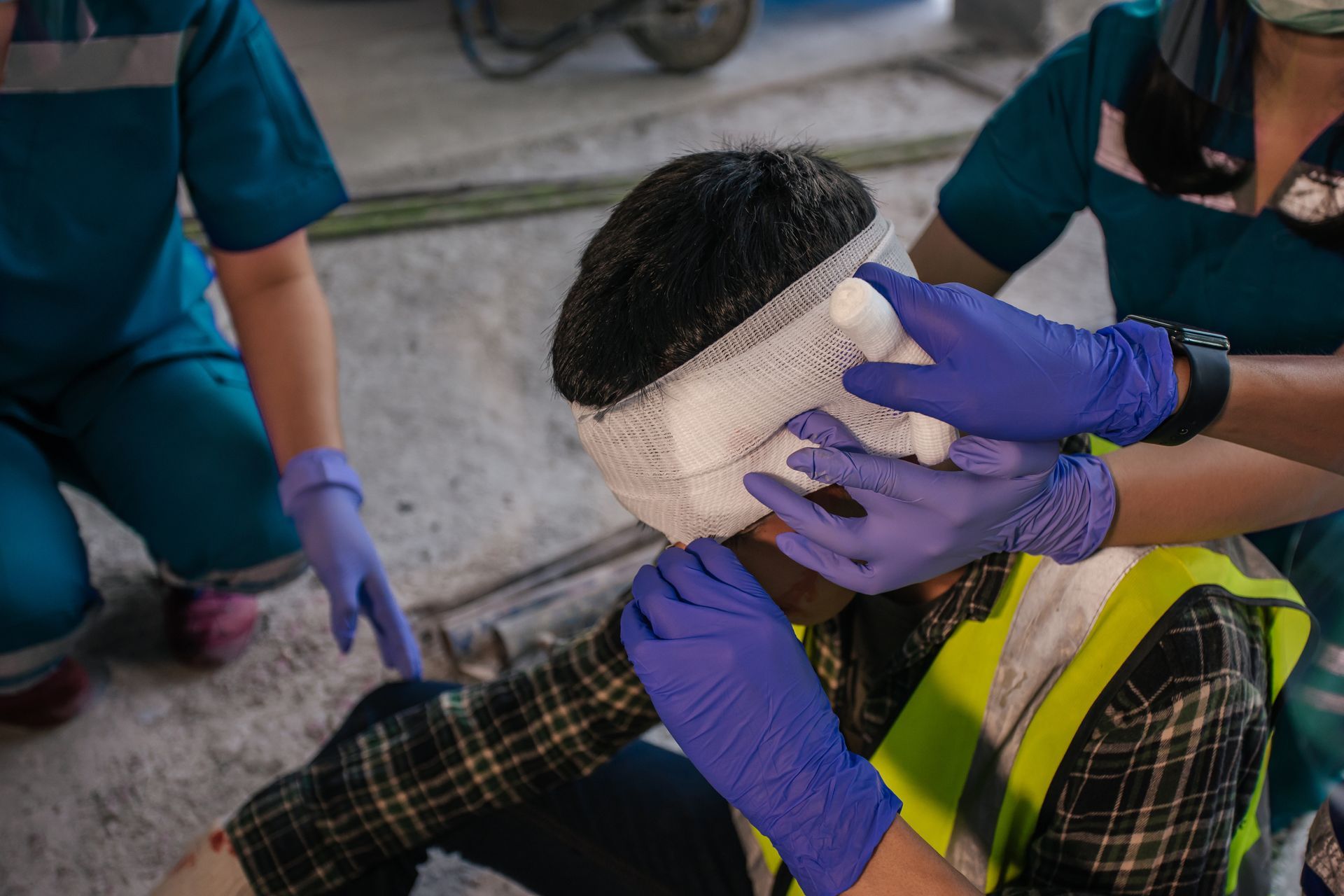 Person in a yellow safety vest receiving a head bandage from gloved responders indoors