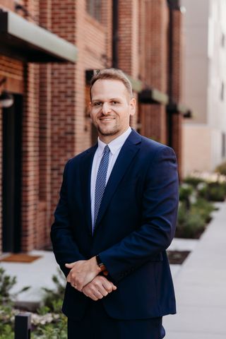 Smiling person in a gray suit and tie standing by windows in a bright indoor setting