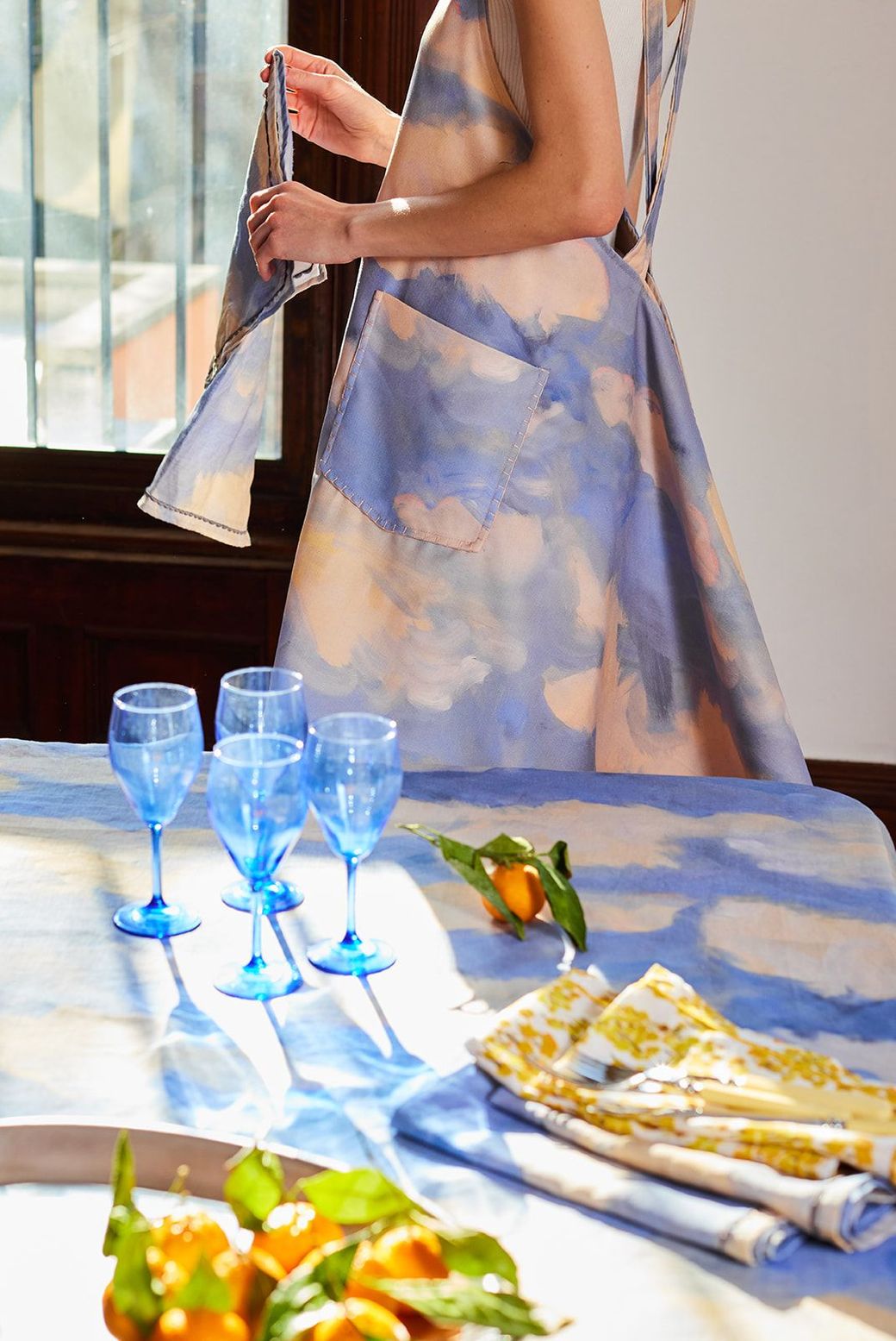 A woman in a blue dress is standing next to a table with blue wine glasses and oranges.