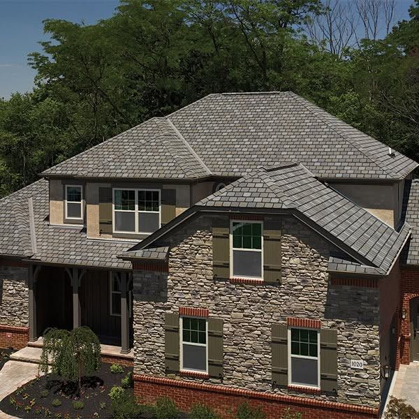 Two-story house with gray stone siding and a shingled roof, viewed from above amid trees