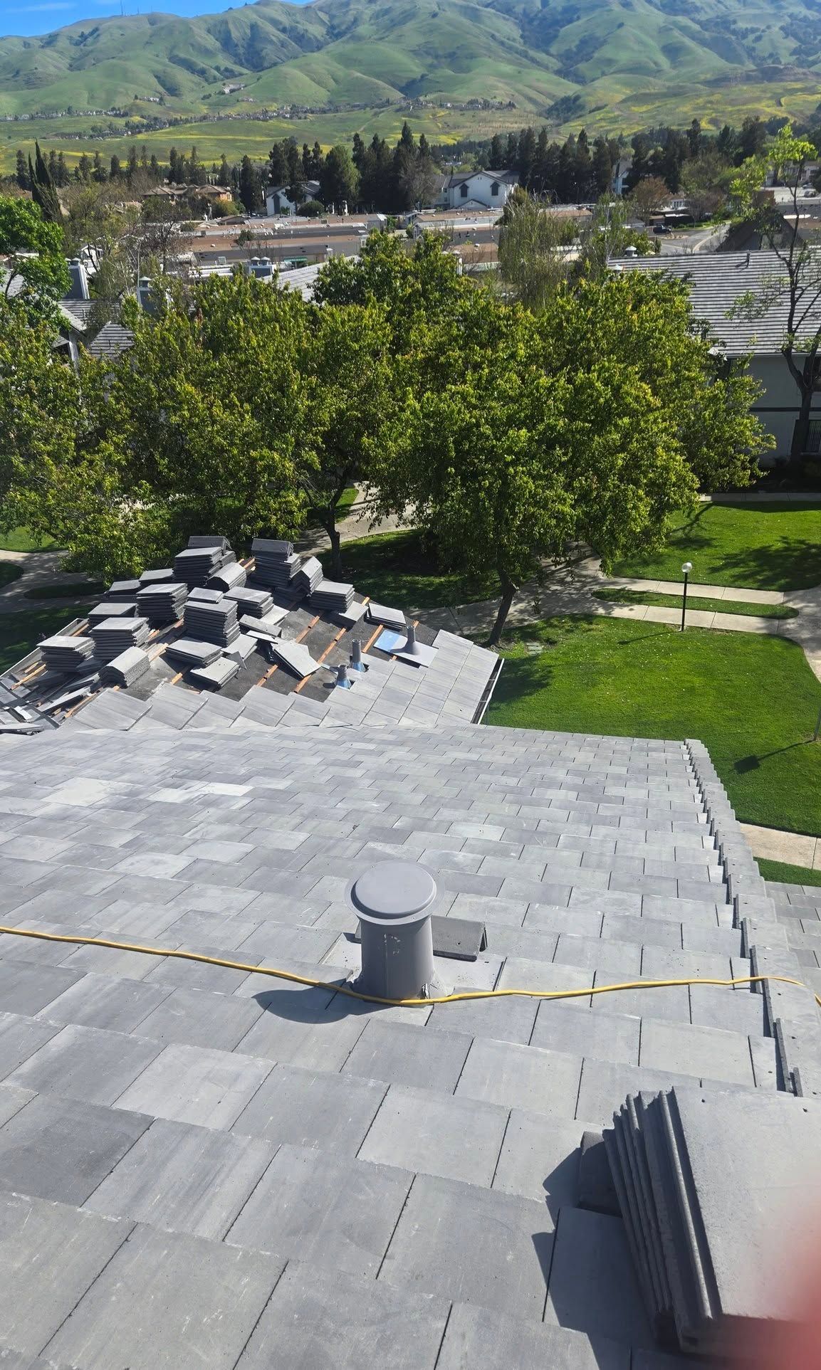 Rooftop with gray shingles, vents, and trees overlooking a town and green mountains under a clear sky