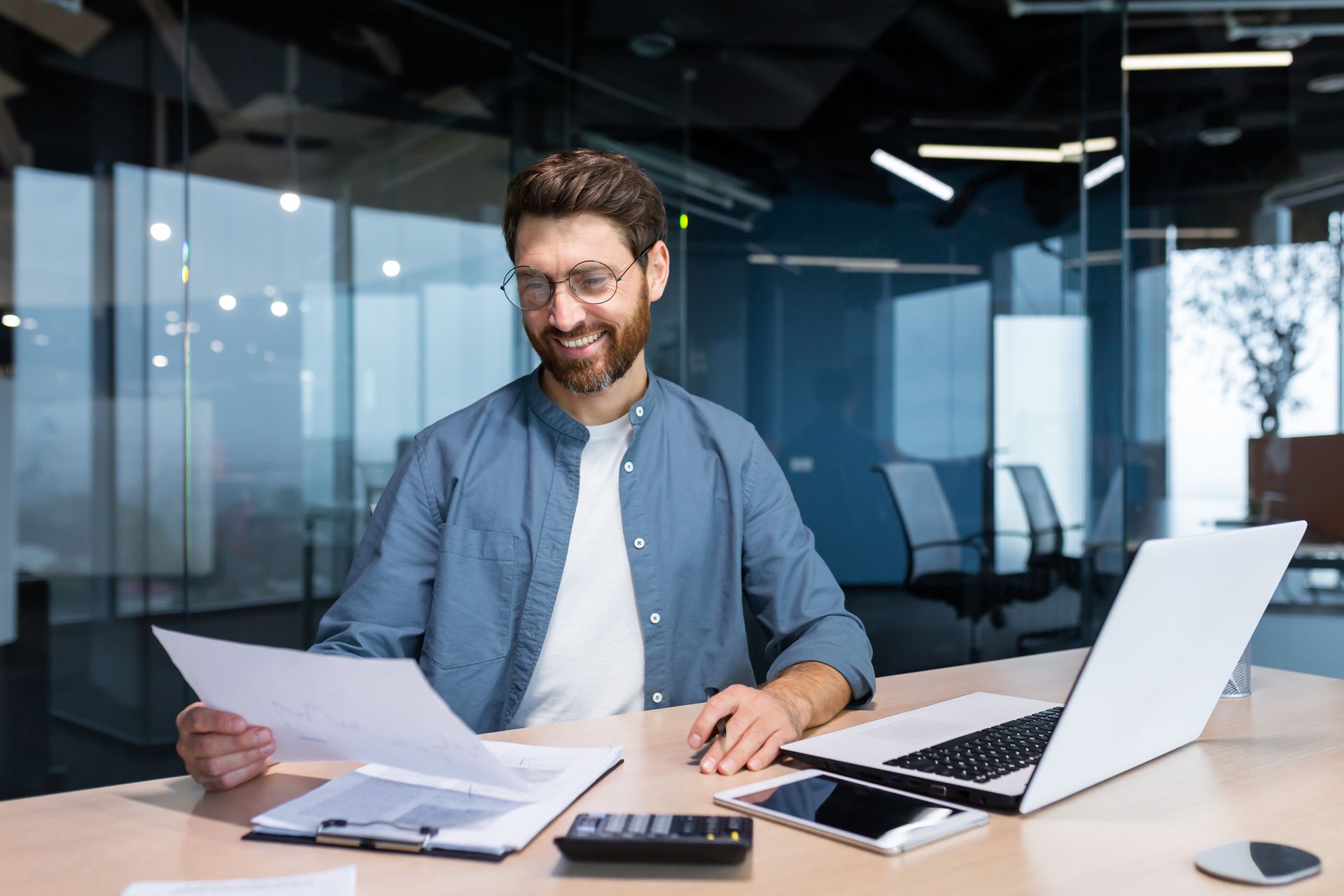 Man with beard smiling, reviewing documents at desk with laptop, calculator, and phone in modern office.