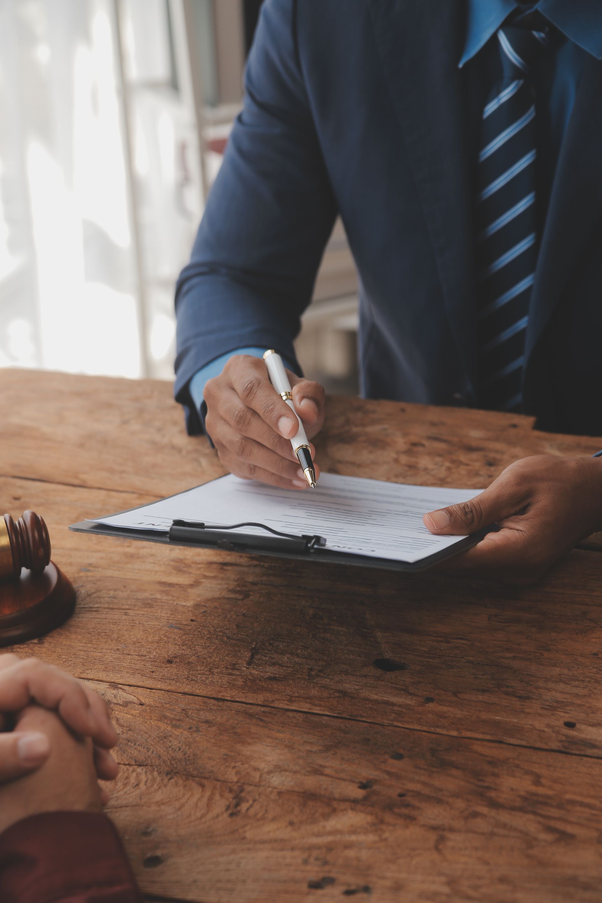 Person in suit signing document on a clipboard at a wooden table. A gavel rests nearby.