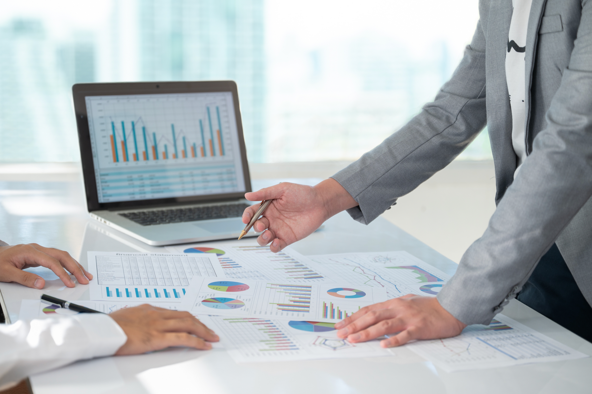 Two people reviewing charts and graphs on a laptop and printed papers at a white table in an office setting.