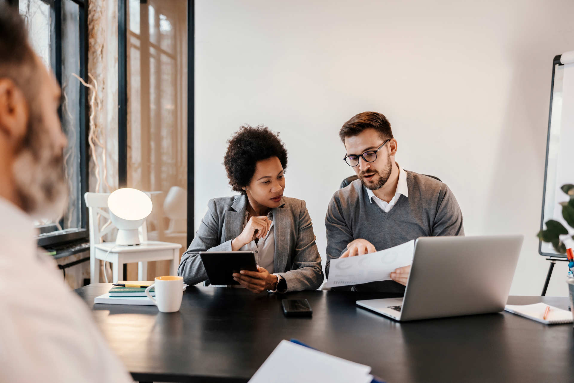 Business colleagues reviewing documents at a table. Woman holds tablet; man gestures at papers. Light streams through window.