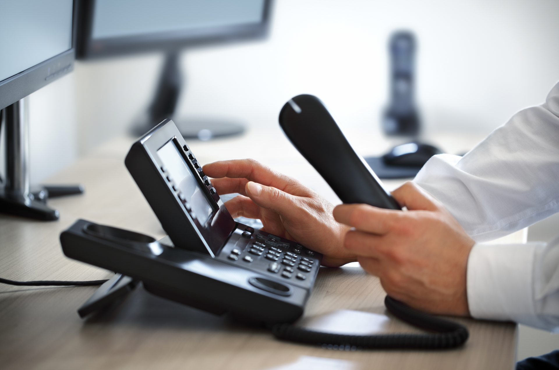 Person using a desk phone, holding handset, with computer monitors in the background.
