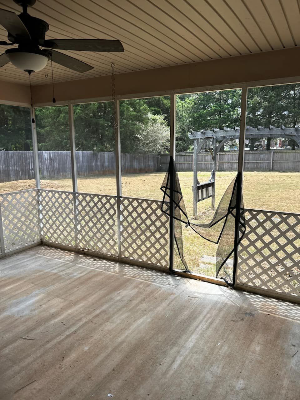A screened-in porch with a wooden floor, a ceiling fan, white lattice railings, and a torn screen door leading to a yard.