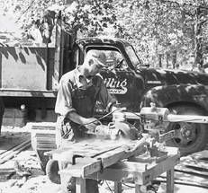 Vintage shot of Kreiling roofer masterfully cutting lumber with a radial arm saw, high lift truck in backdrop Vintage shot of Kreiling roofer masterfully cutting lumber with a radial arm saw, high lift truck in backdrop