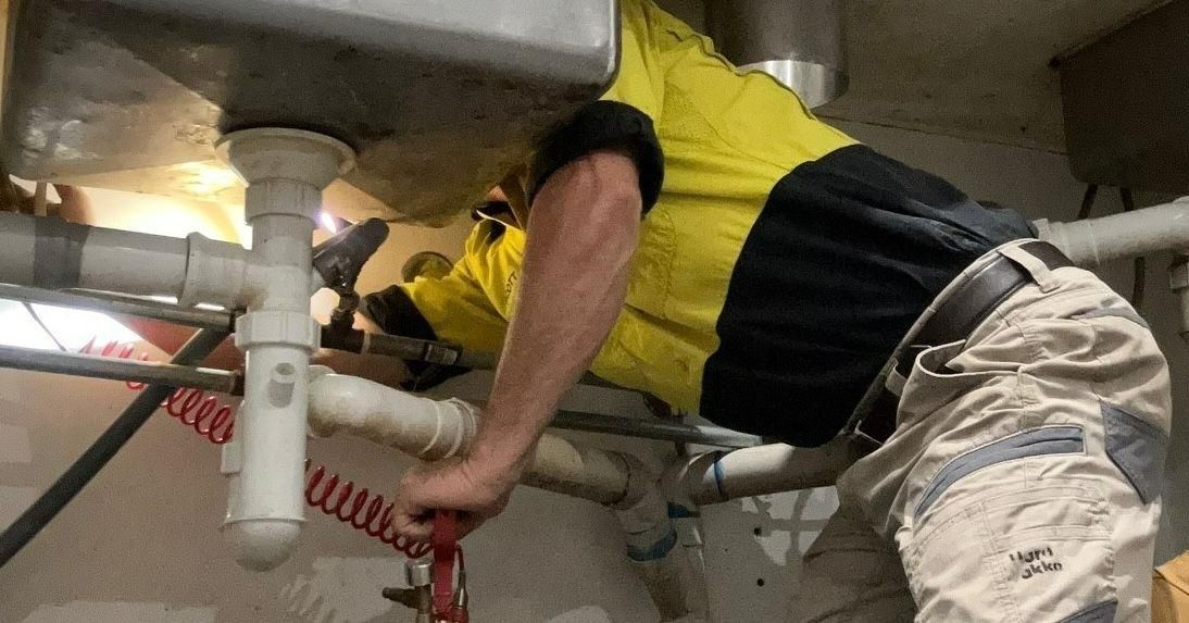 A Man in a Yellow Shirt is Fixing a Sink With a Wrench — Allied Pest Management Northern Rivers in Evans Head, NSW