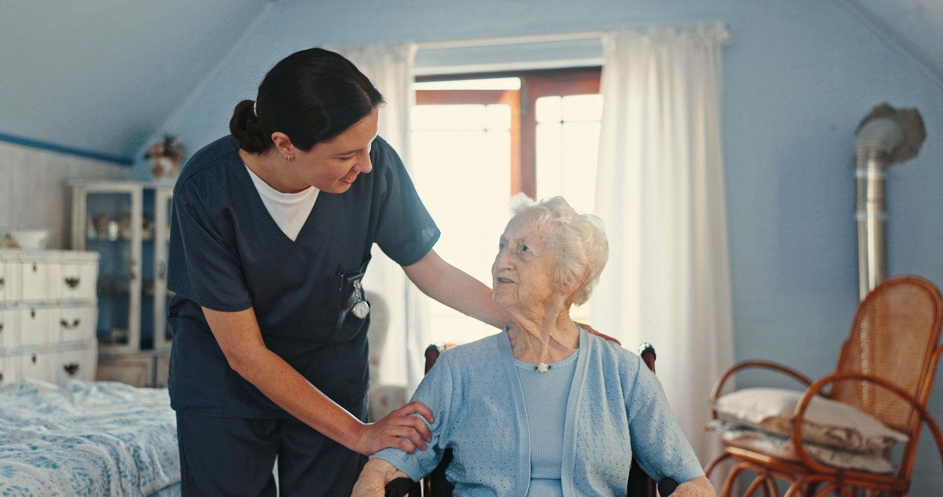 Caregiver smiling and holding hands with an elderly woman in a bright bedroom