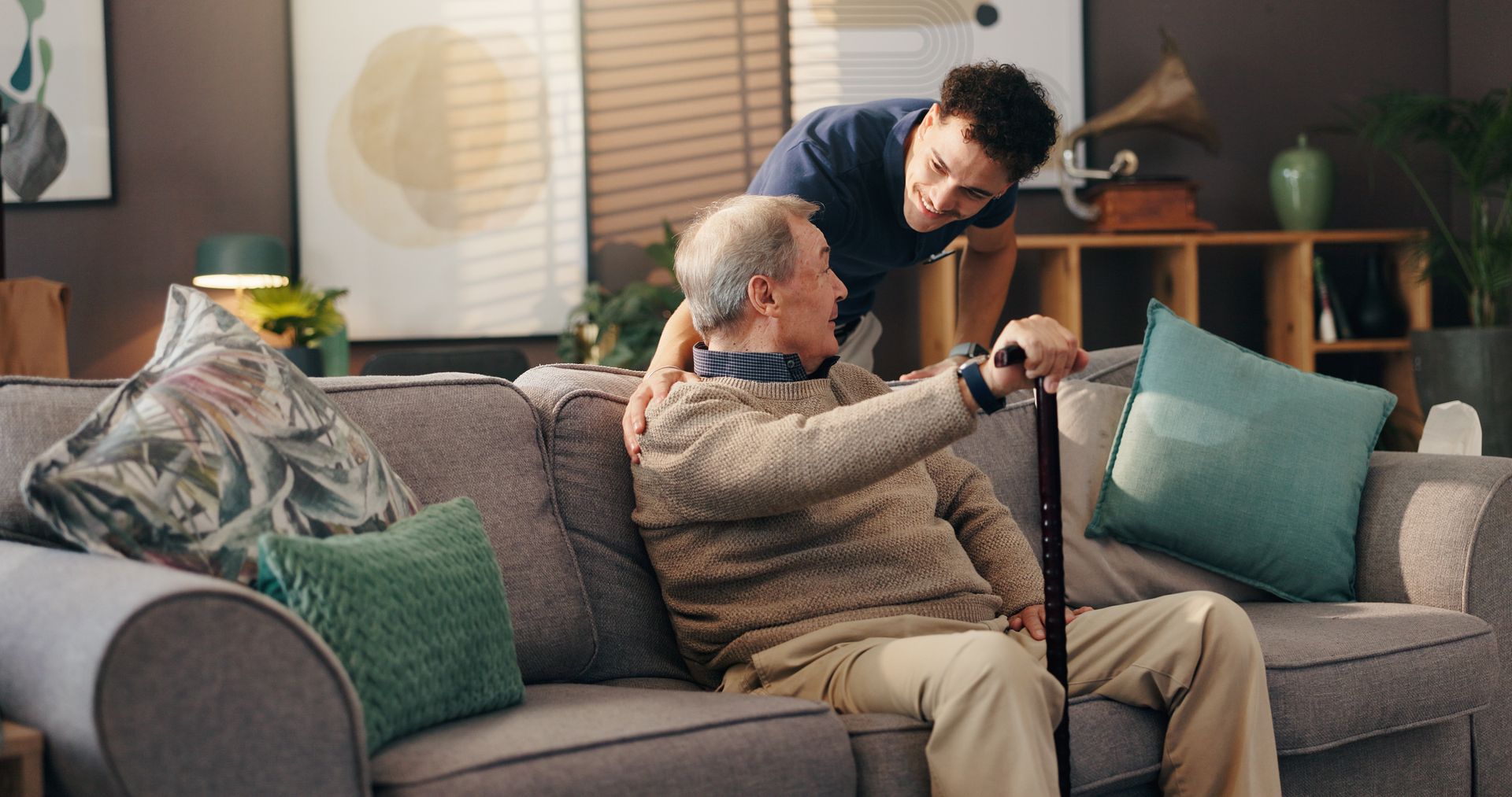 Two people on a sofa, one sitting with a cane while the other helps adjust a device in a living room