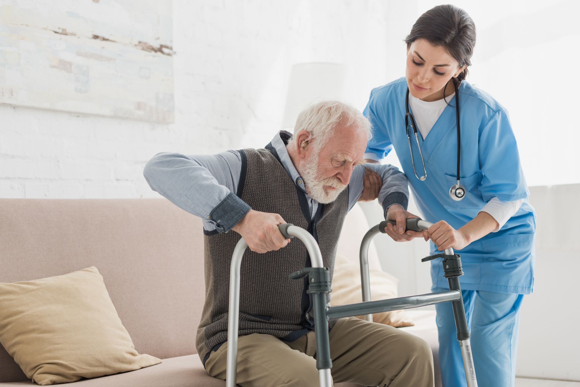 Caregiver in blue scrubs helps an older man stand with a walker in a bright living room
