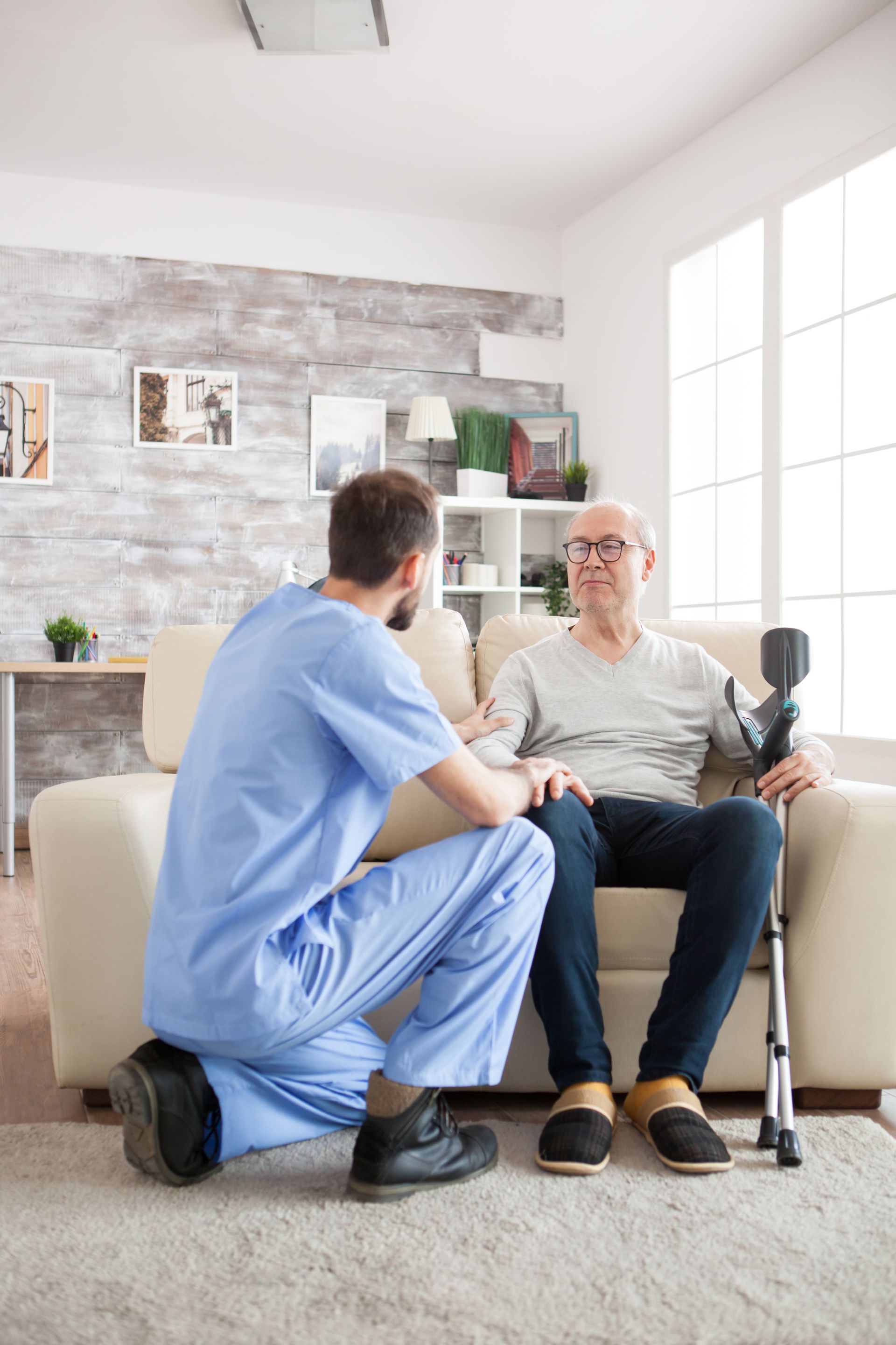 Caregiver kneels beside seated patient in a bright living room, speaking and holding the patient's arm.