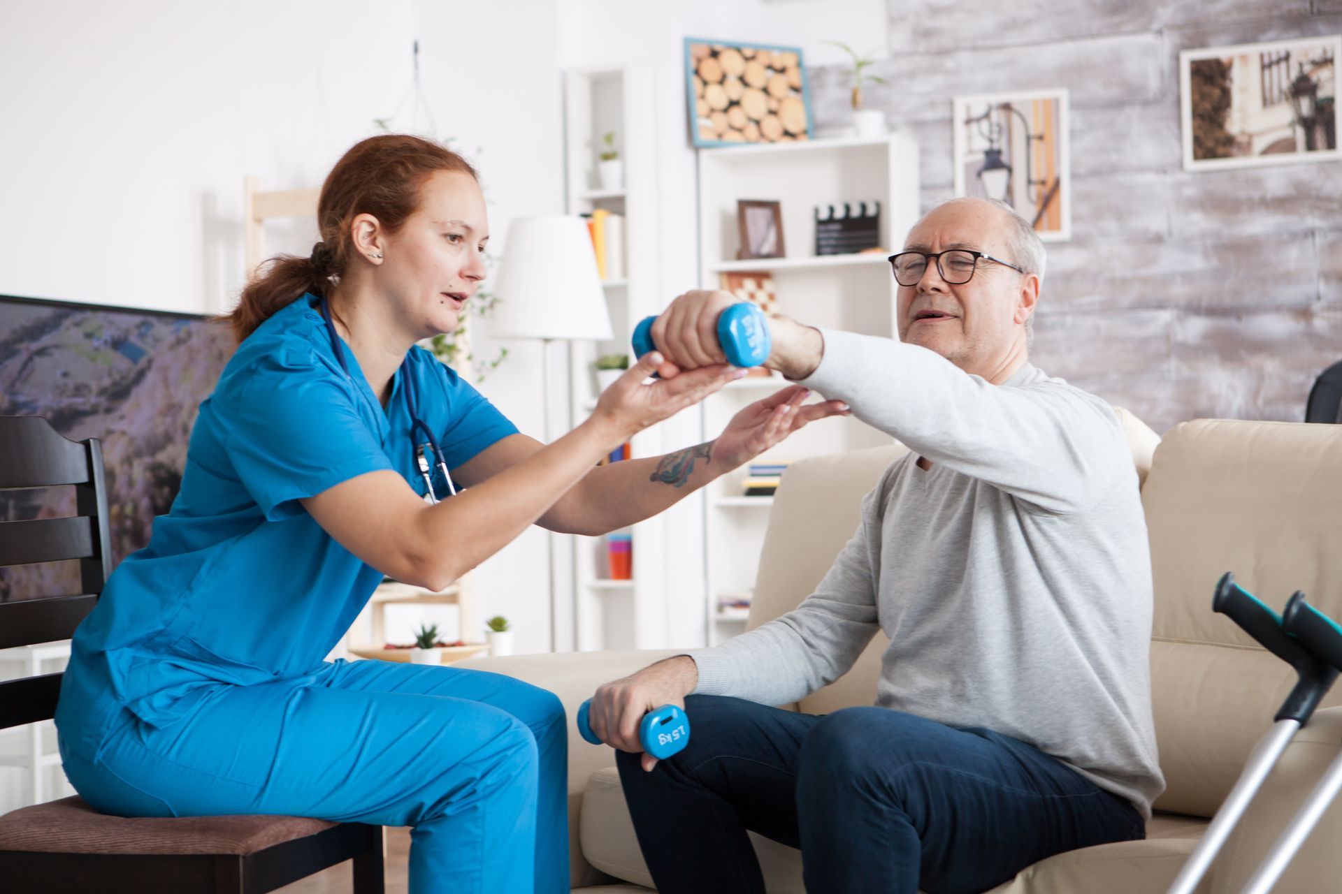 Caregiver in blue helping seated man use a dumbbell in a bright living room.