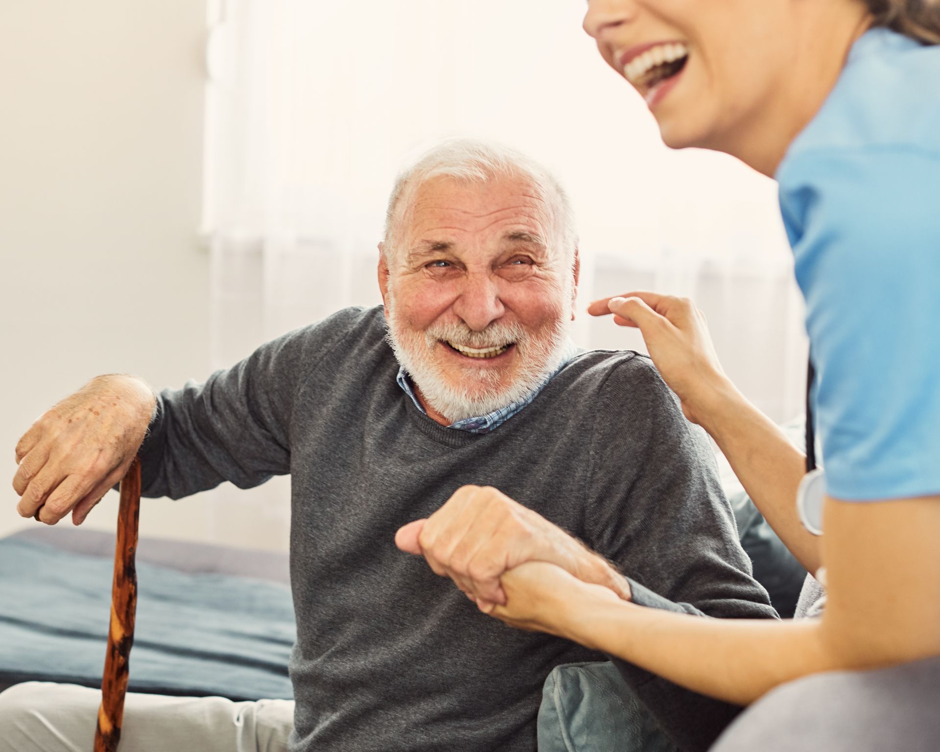 Smiling elderly man with a cane sits beside a caregiver in a bright bedroom